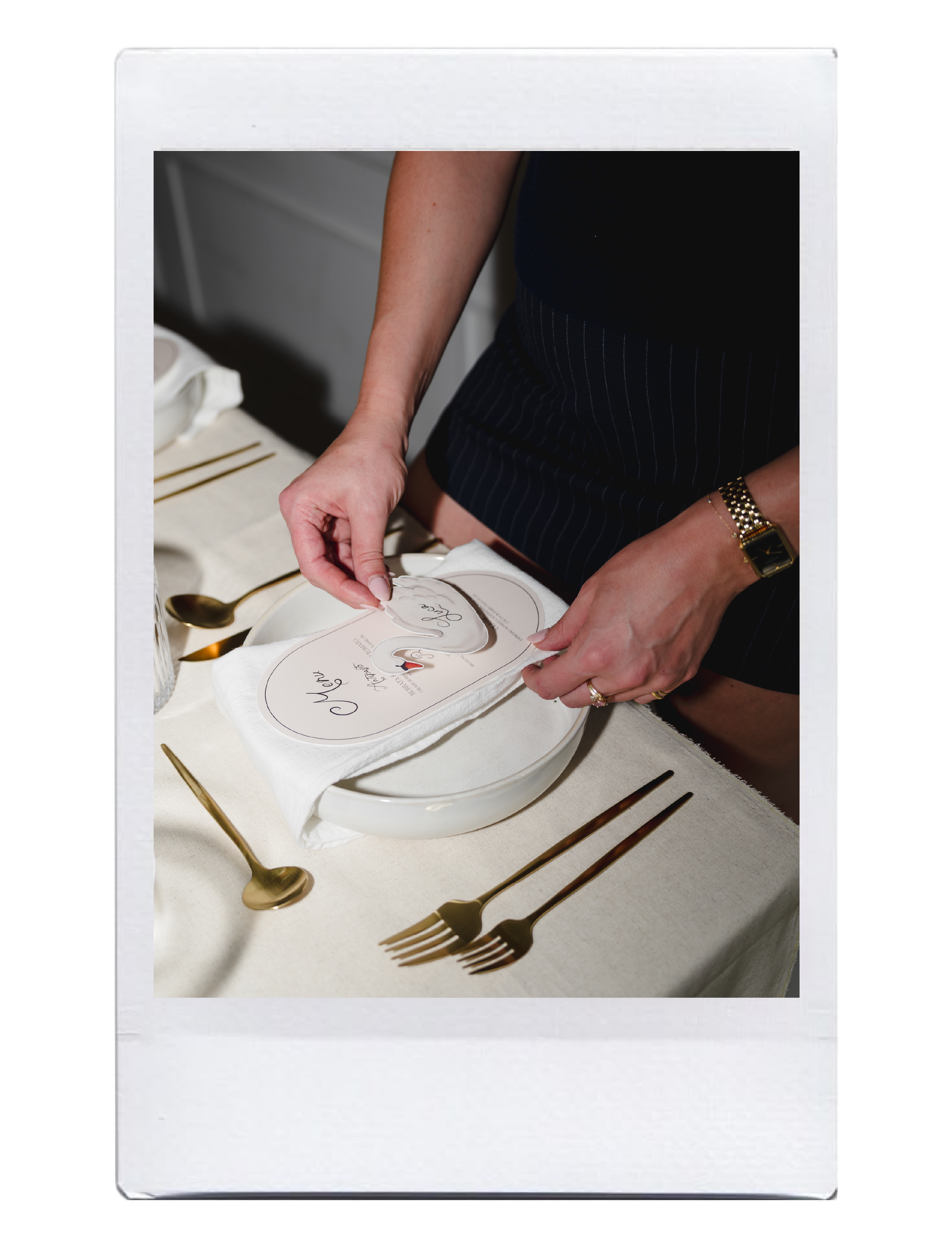 A person is wrapping a white towel with a heart-shaped card on top of a gift box. The person is wearing a gold wristwatch and a dark top. The gifts are placed on a beige tablecloth, with three gold-colored forks arranged in a row.