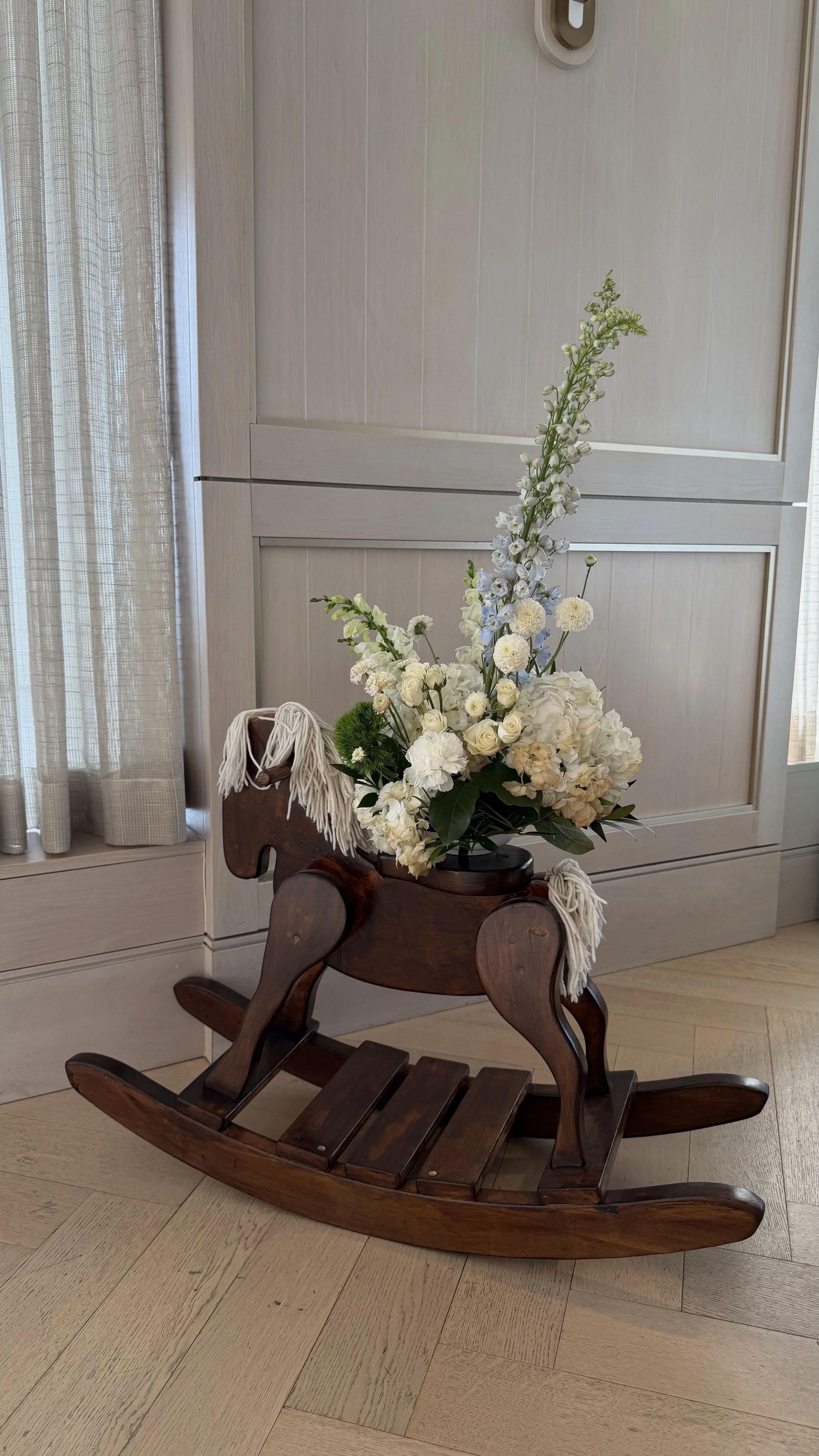 A wooden rocking horse with a bouquet of white flowers on it, placed in front of a cabinet and near a window with curtains.