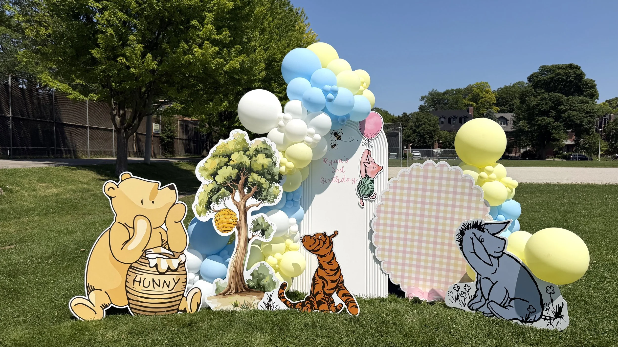 Decorations for a child's birthday party featuring cartoon animal cutouts of a bear labeled 'Hunny', a tiger, and an elephant, with colorful balloons and a sign reading 'Ryan's 3rd Birthday' in a park or playground area.