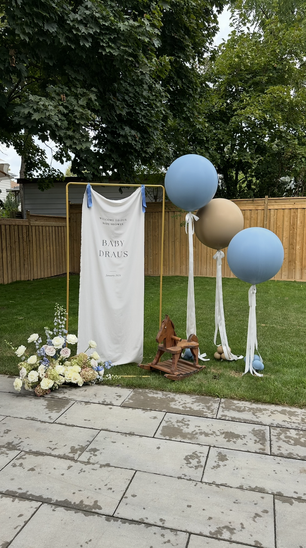 Decorations for a baby shower with a sign that reads "Welcome to Our Baby Shower, Baby Draus, January 2023." There are three large balloons, two blue and one beige, tied with white ribbons. A small wooden rocking horse is on the grass, and a floral a