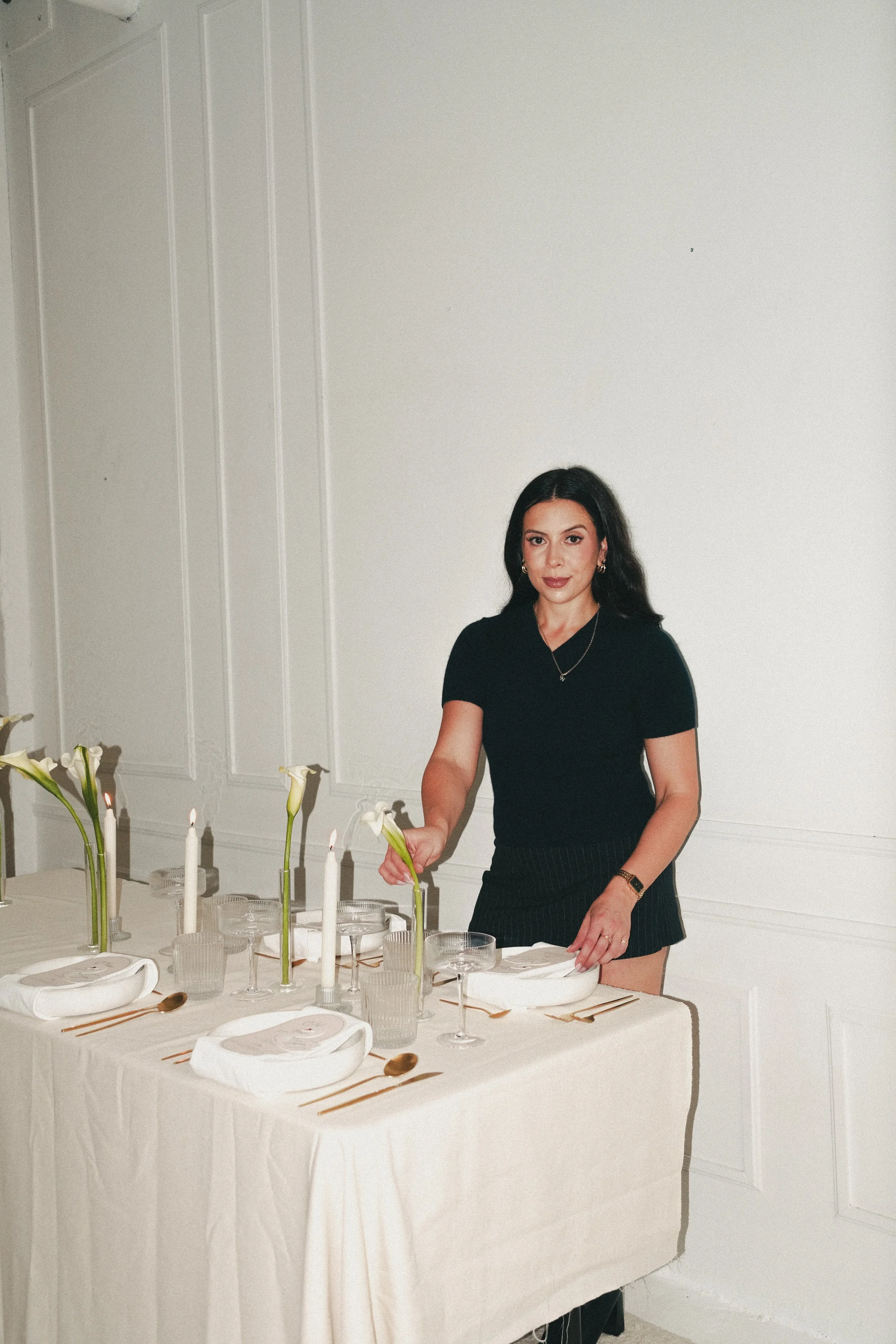 A woman with long black hair, wearing a black dress and jewelry, standing beside a dining table with white tablecloth, modern white candles, flowers, and gold flatware, in a minimalist room with white walls and decorative paneling.