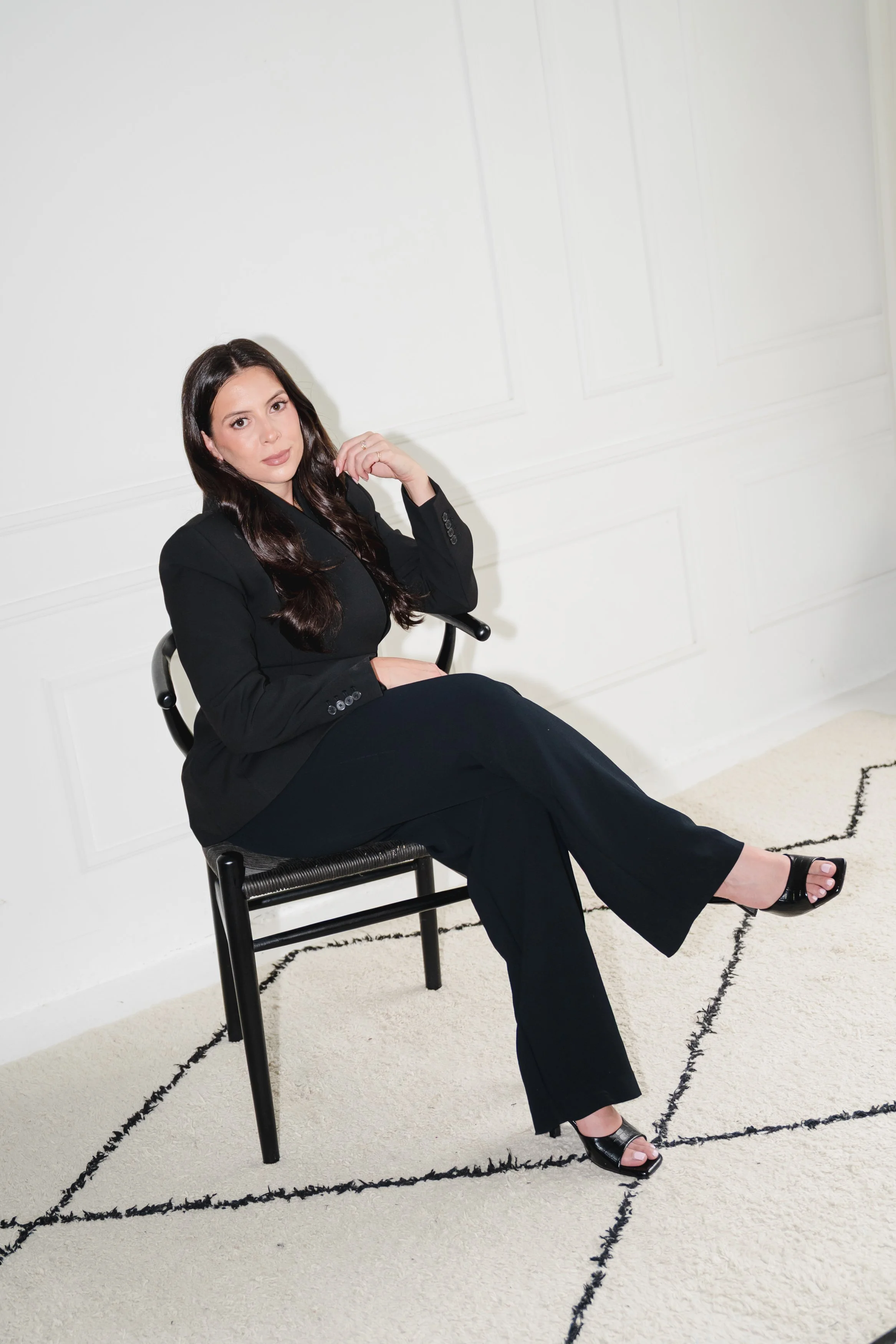 A woman with long dark hair sitting on a black chair in a white room, wearing a black suit and open-toe black heels, with her left leg crossed over her right and her right hand resting on her thigh.