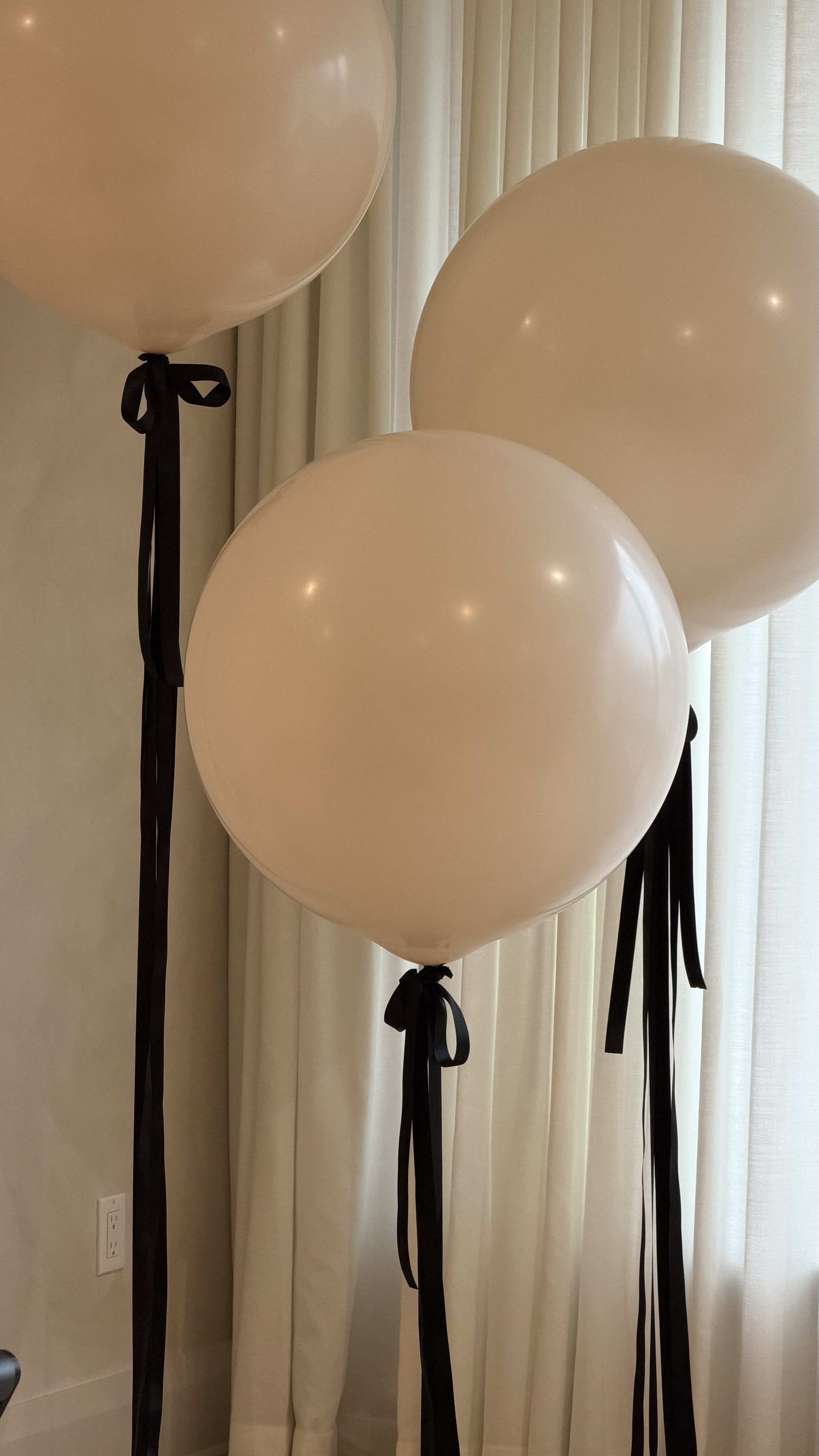 Five white balloons tied with black ribbons, arranged on black stands, set against cream curtains.