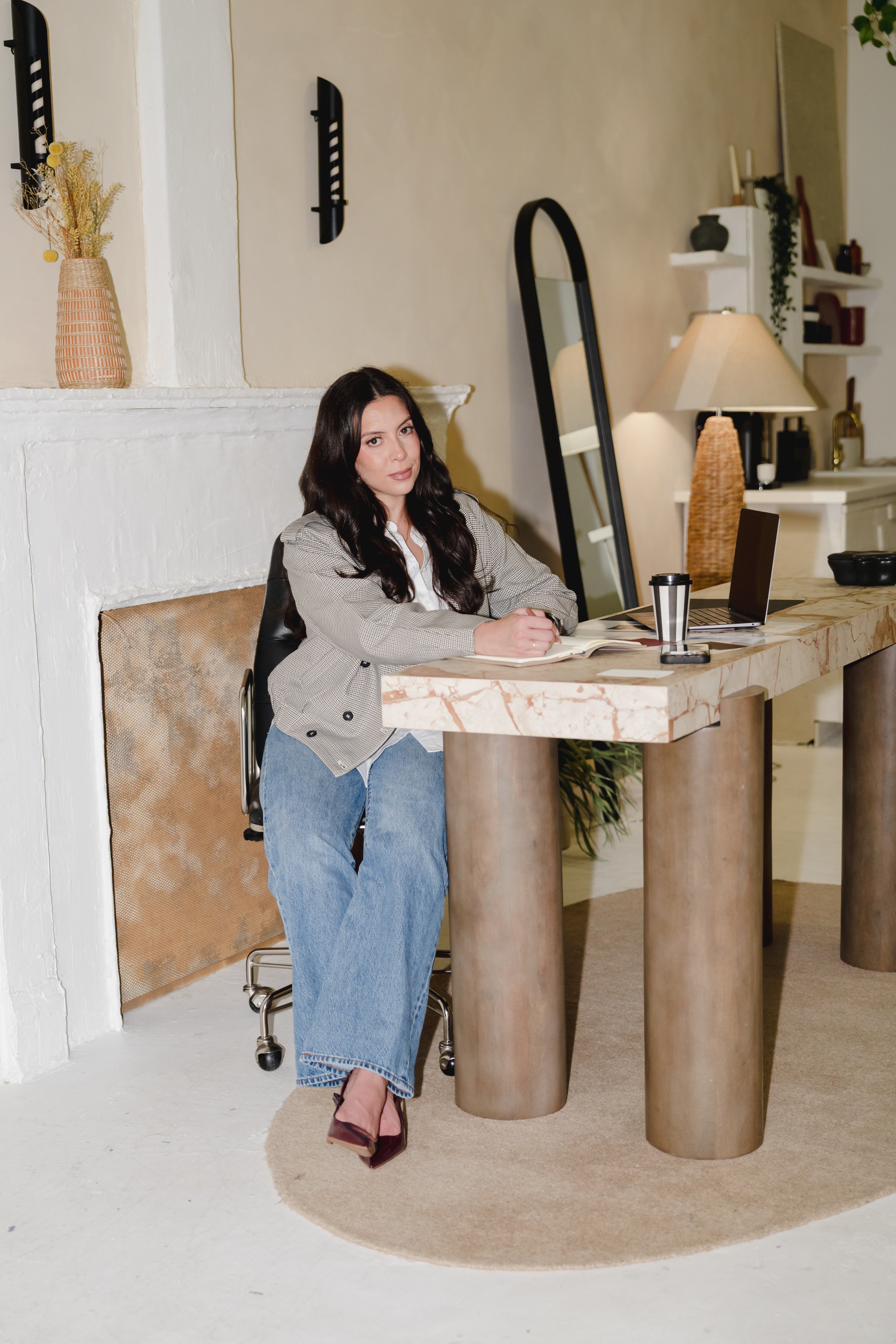 A woman with long dark hair sitting behind a marble-topped table in a cozy room, with a laptop, notebook, and coffee cup in front of her. The room is decorated with a table lamp, a tall mirror, and white shelves with decorative items and plants.