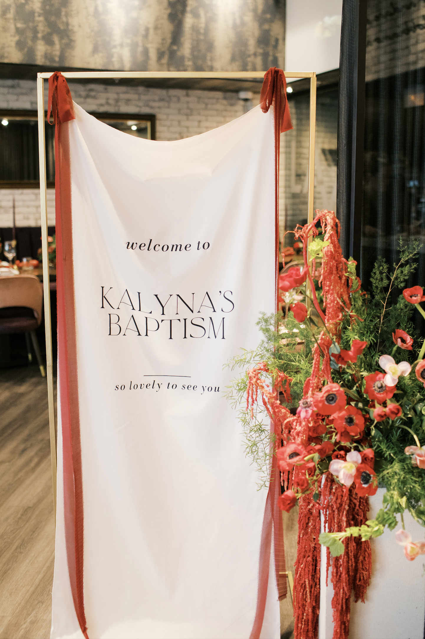 A welcome sign for Kalyna's baptism surrounded by red floral arrangements and ribbons inside a venue with brick walls and wooden floors.