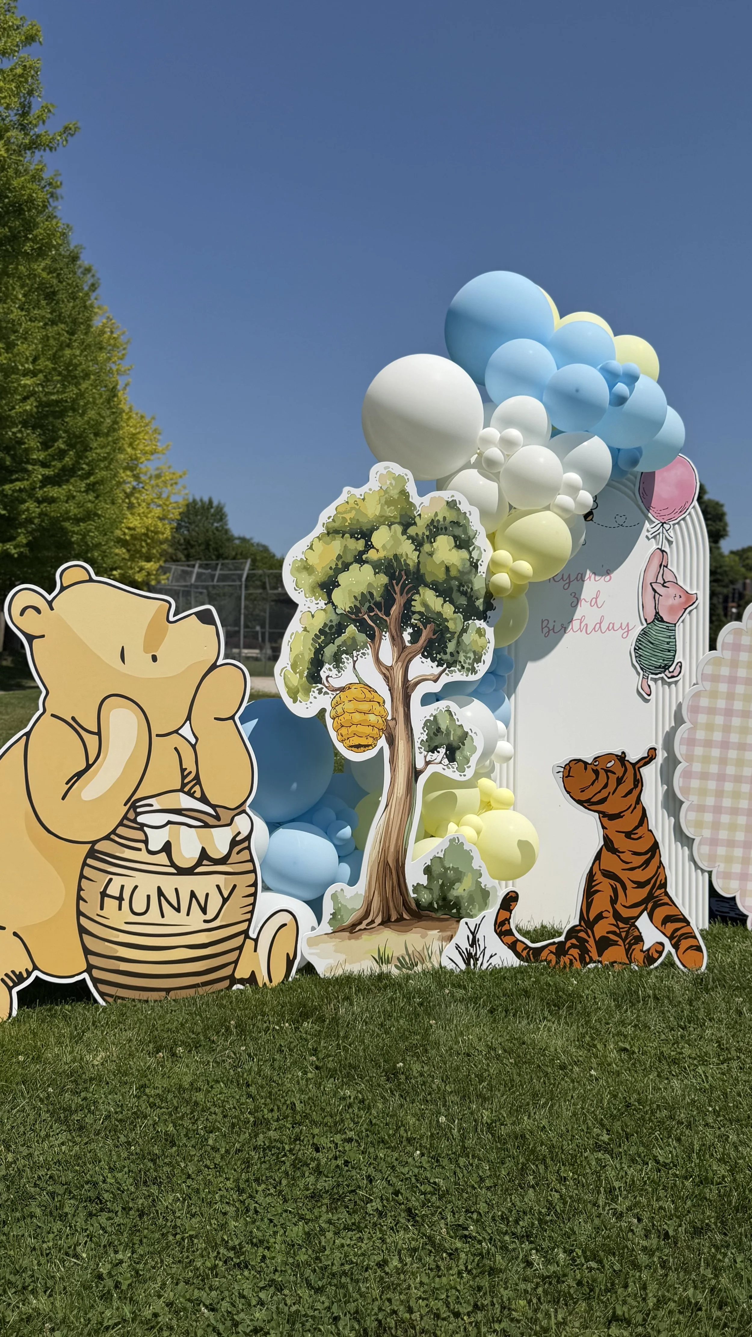 Decorative birthday display featuring cartoon animals including Winnie the Pooh holding honey, a tiger, and pig in a party hat, with a tree and balloons, set outdoors on a grassy area.