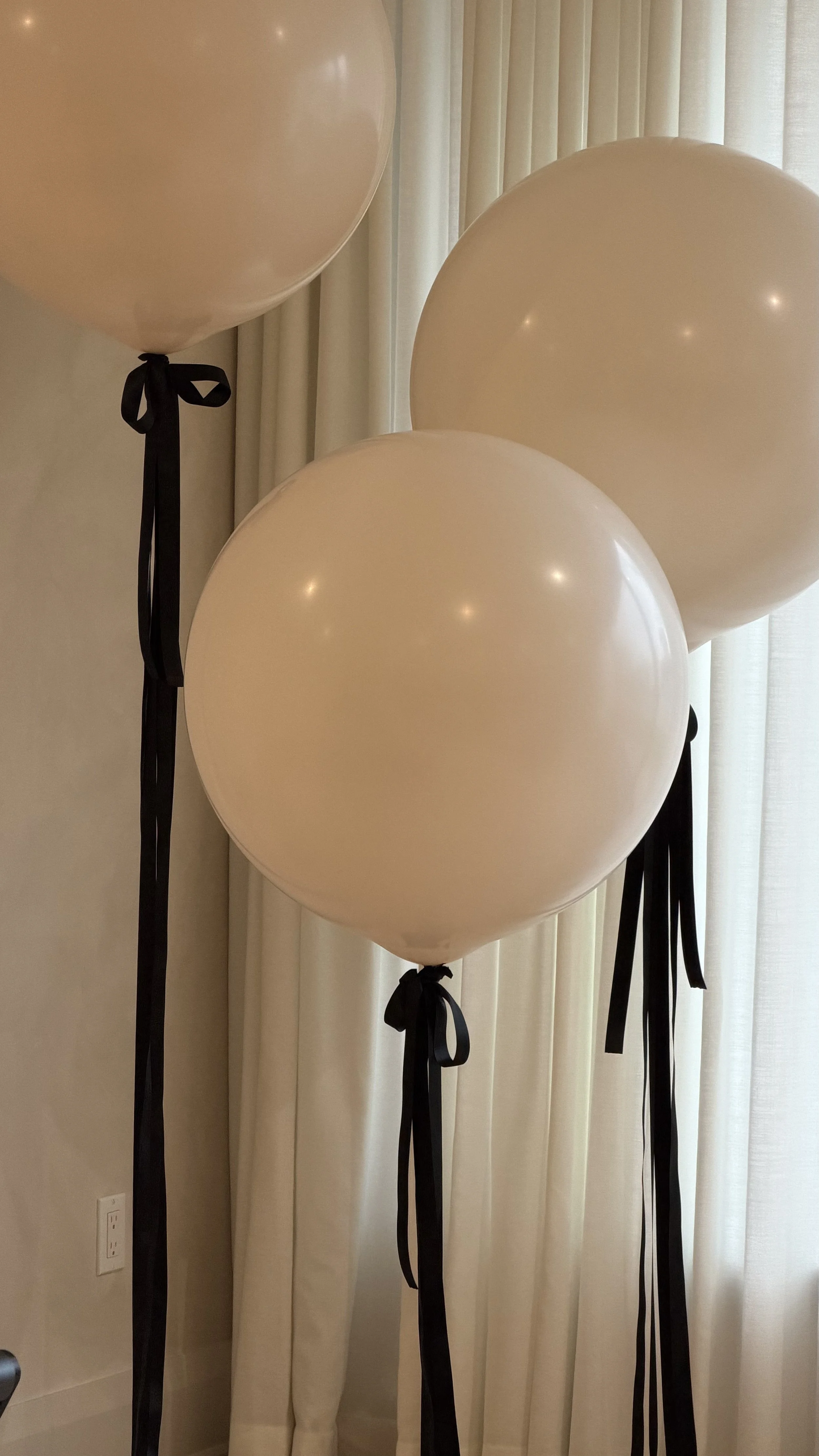 Three beige balloons tied with black ribbons, attached to black strings, standing in front of cream-colored curtains.