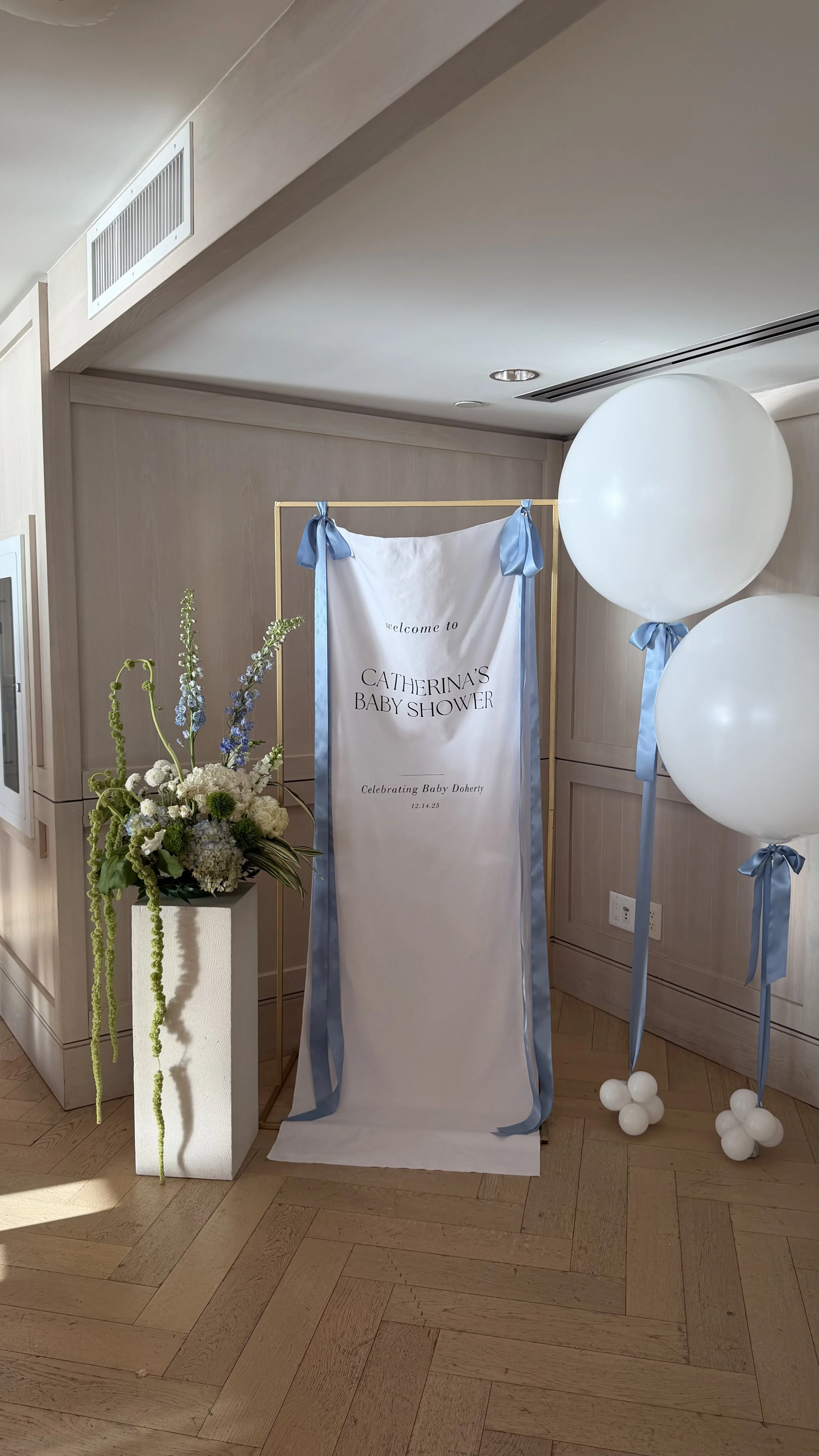 Decorative display for Catherina's baby shower includes a white fabric backdrop with blue ribbons, large white balloons tied with blue ribbons, a floral arrangement with white and purple flowers, and a wooden floor.