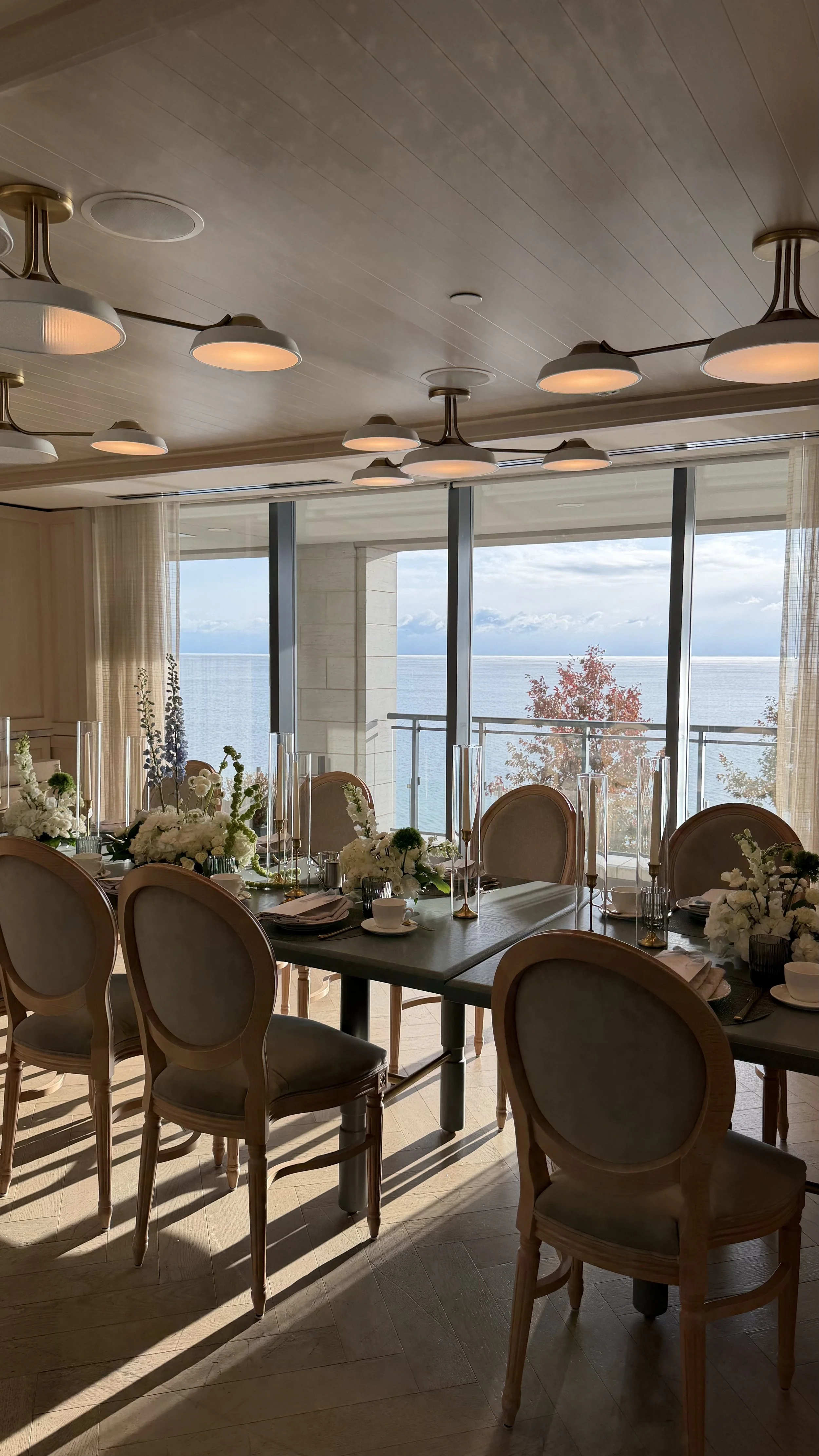 Elegant dining room with round backed chairs, a long table set with white flowers and candles, set for a meal, large windows showing a view of the ocean and trees, modern hanging lights on a wooden ceiling
