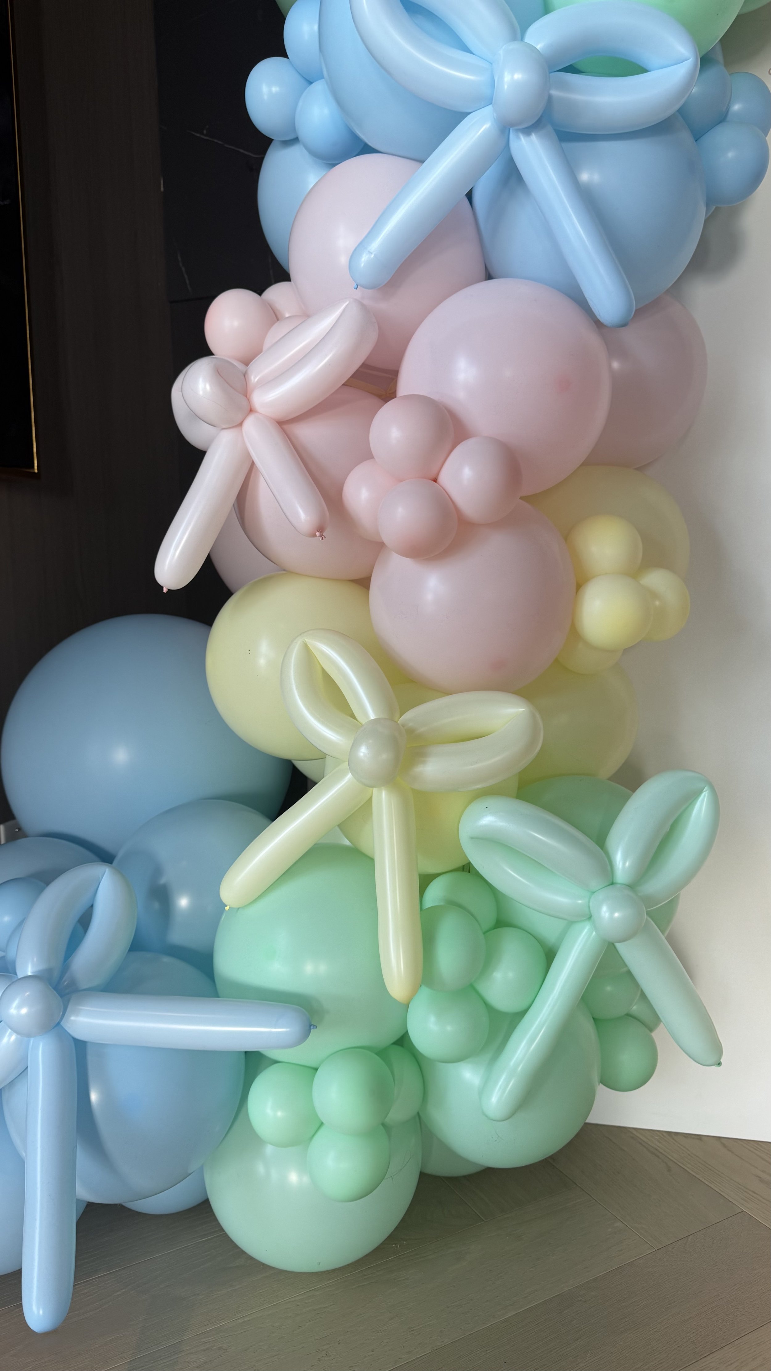 Colorful balloon bouquet with pink, blue, green, and yellow balloons arranged in floral patterns, some shaped like flowers with balloon petals and centers.