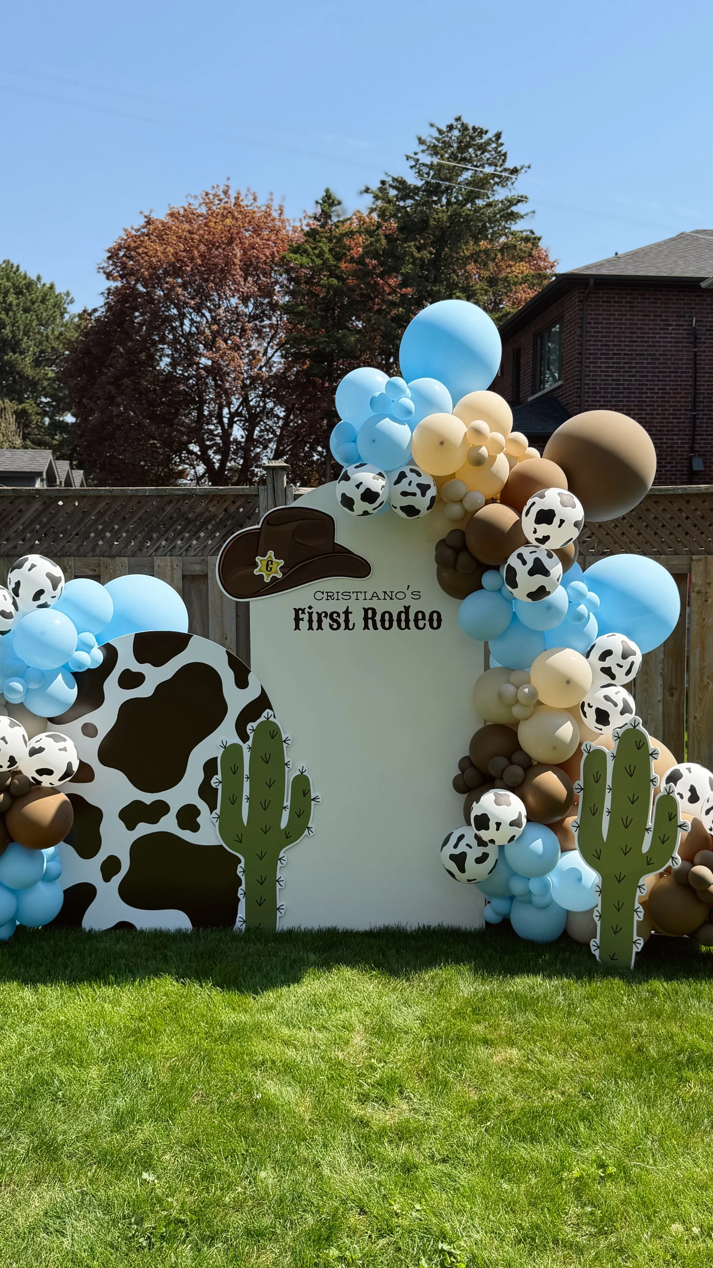 Decorative outdoor setup with balloons, cow print patterns, and cacti for a first rodeo event, featuring a sign that reads "Cristiano's First Rodeo" with a cowboy hat illustration.