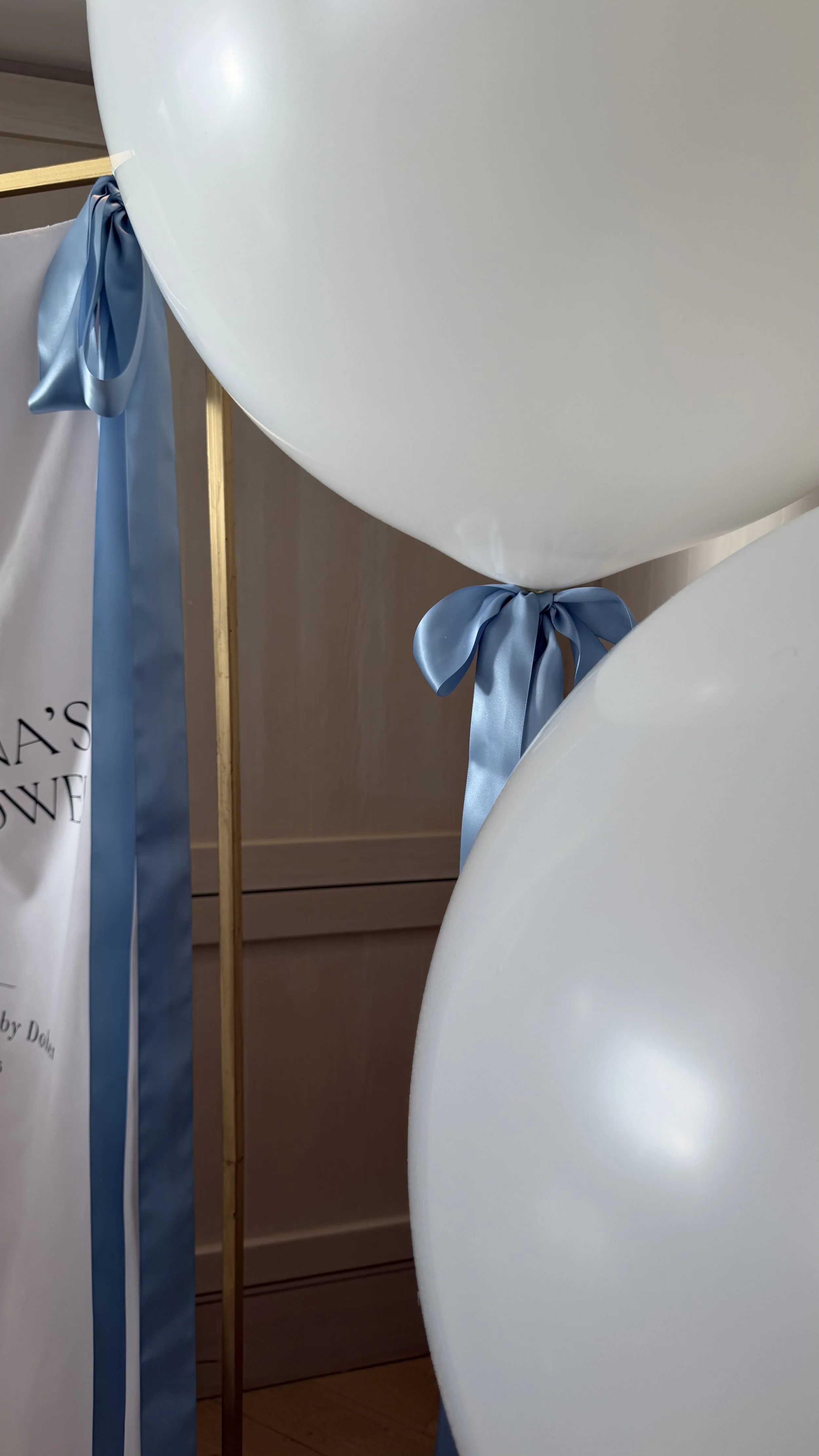 Close-up of white balloons with blue satin ribbons tied around the strings, with a wooden stand and part of a sign in the background.