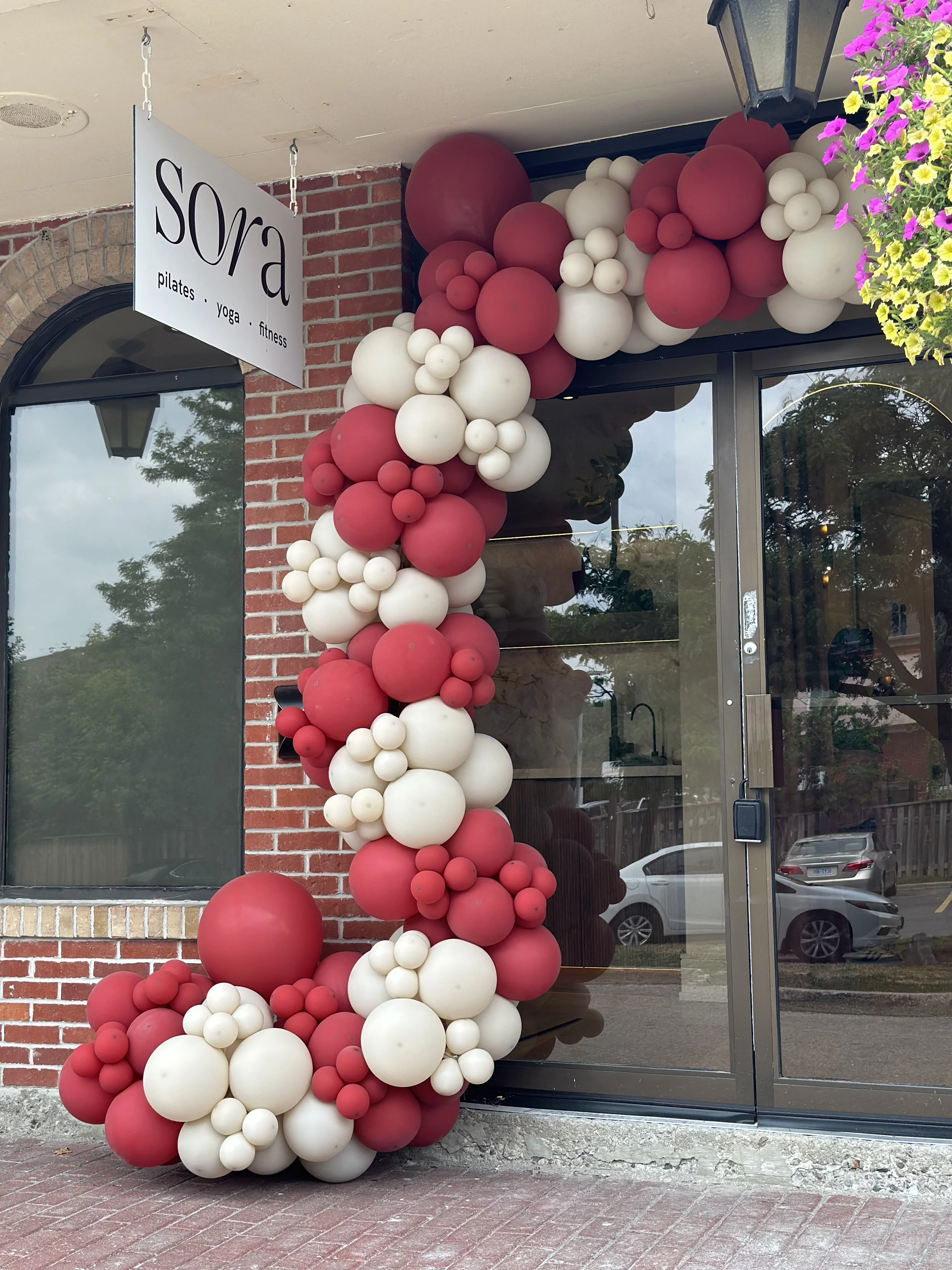 Decorative balloon arrangement in red and white colors outside a storefront with a brick wall and sign reading 'Sora' for Pilates, yoga, and fitness.