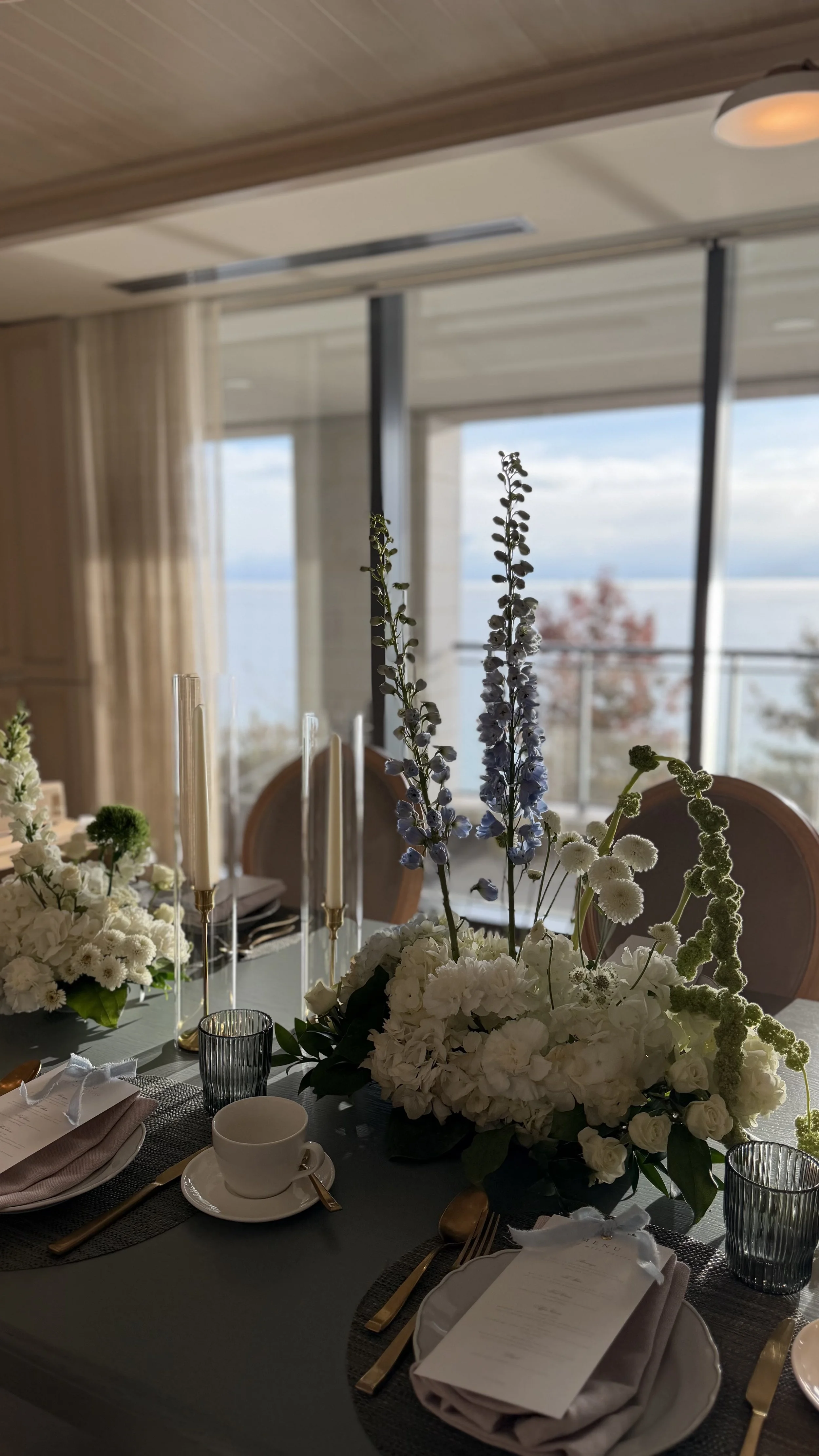 A dining table set for a formal event with a centerpiece of white and purple flowers, surrounded by elegant tableware, and large windows in the background offering a view of the outdoors.