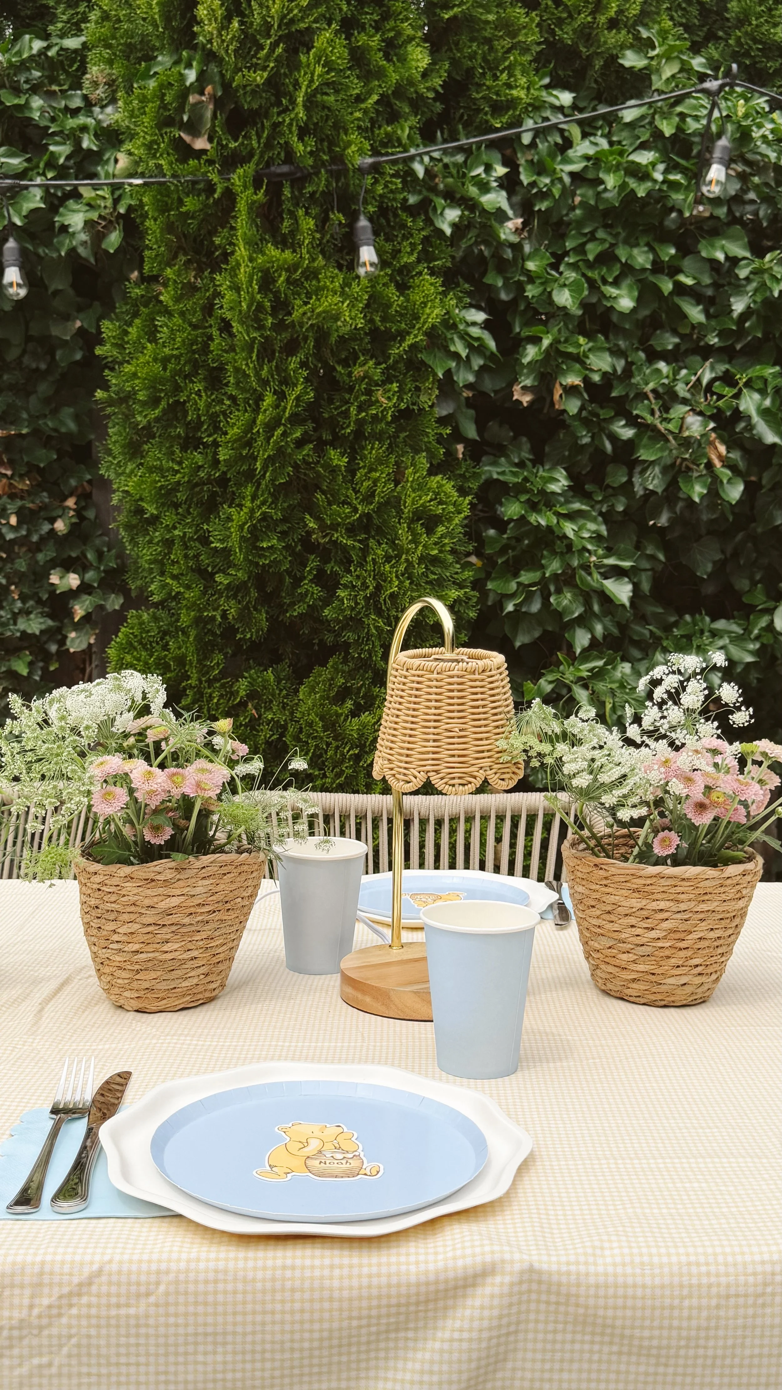 Outdoor dining setup with floral arrangements in baskets, empty cups, and a table lamp, against a backdrop of lush green hedge and trees.