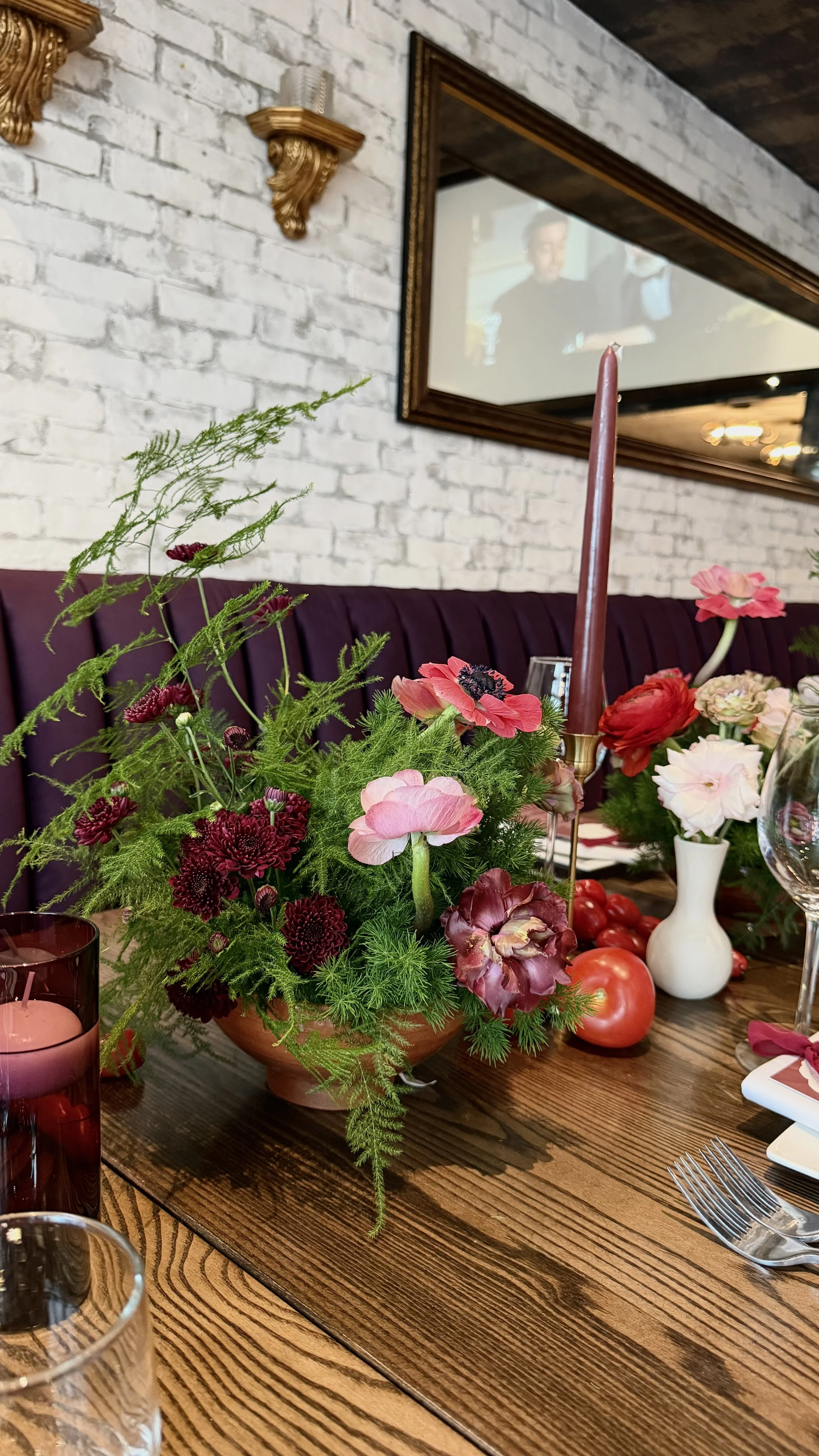A dining table with a colorful floral centerpiece, a pink candle, and some tomatoes. The background features a white brick wall, a mirror, and decorative wall sconces.