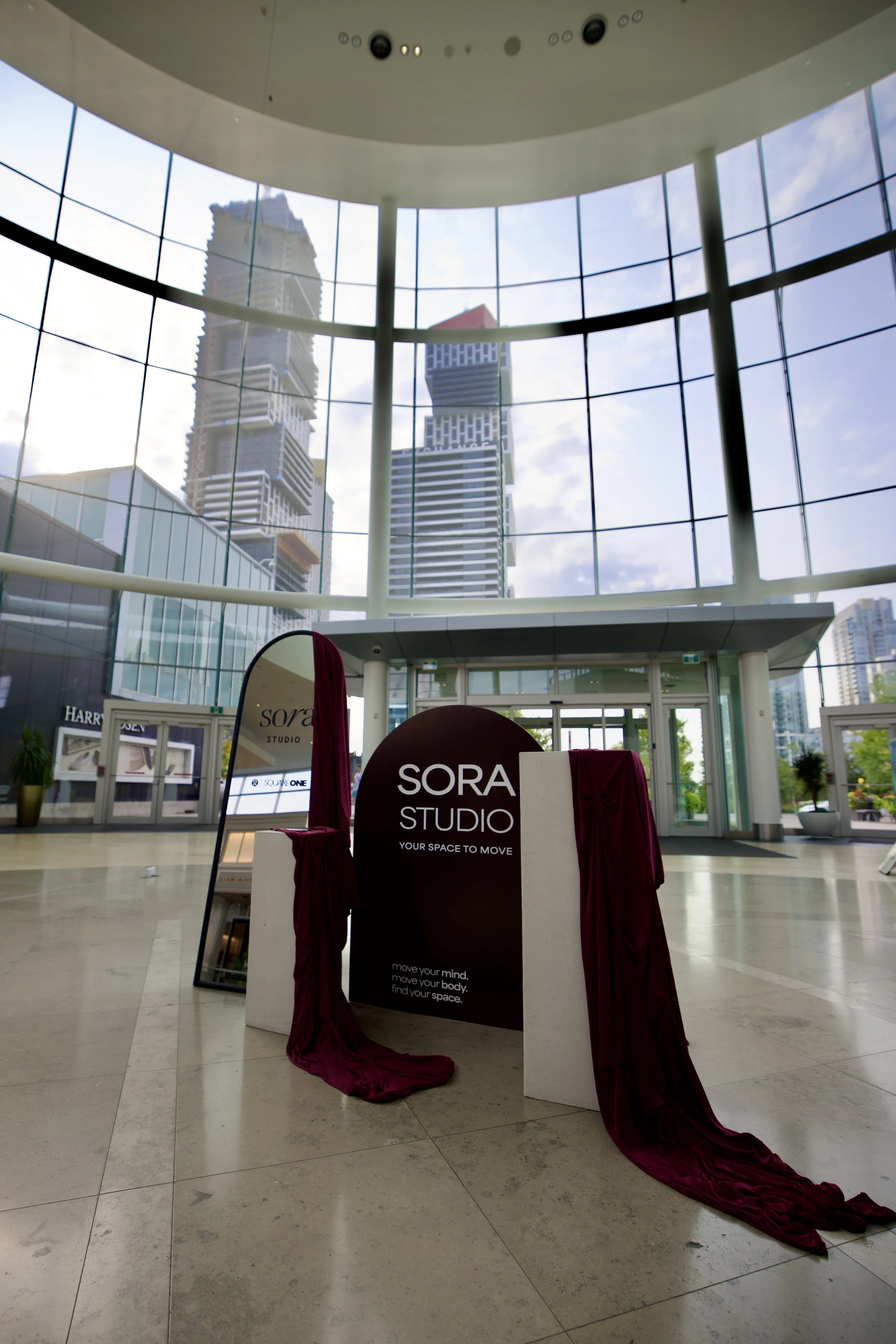 Indoor shopping mall with large glass windows showing tall buildings outside. In the foreground, a sign for SORA STUDIO with the tagline 'Your space to move,' decorated with burgundy fabric draped around it. There are promotional signs and store wind