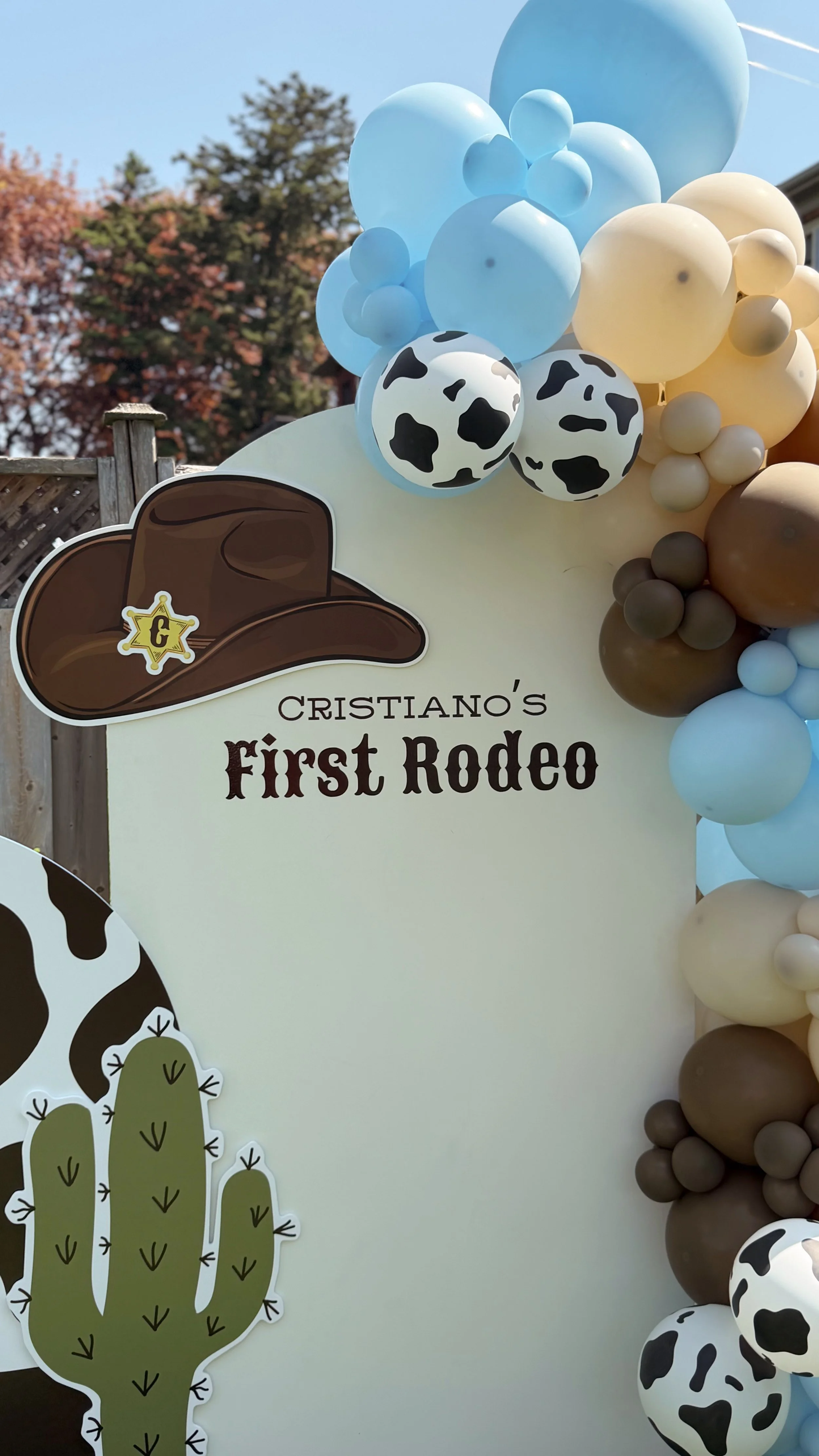 Decorative sign for Cristoano's First Rodeo with cow print balloons, a cow hat graphic, and cactus cutouts in a western theme.