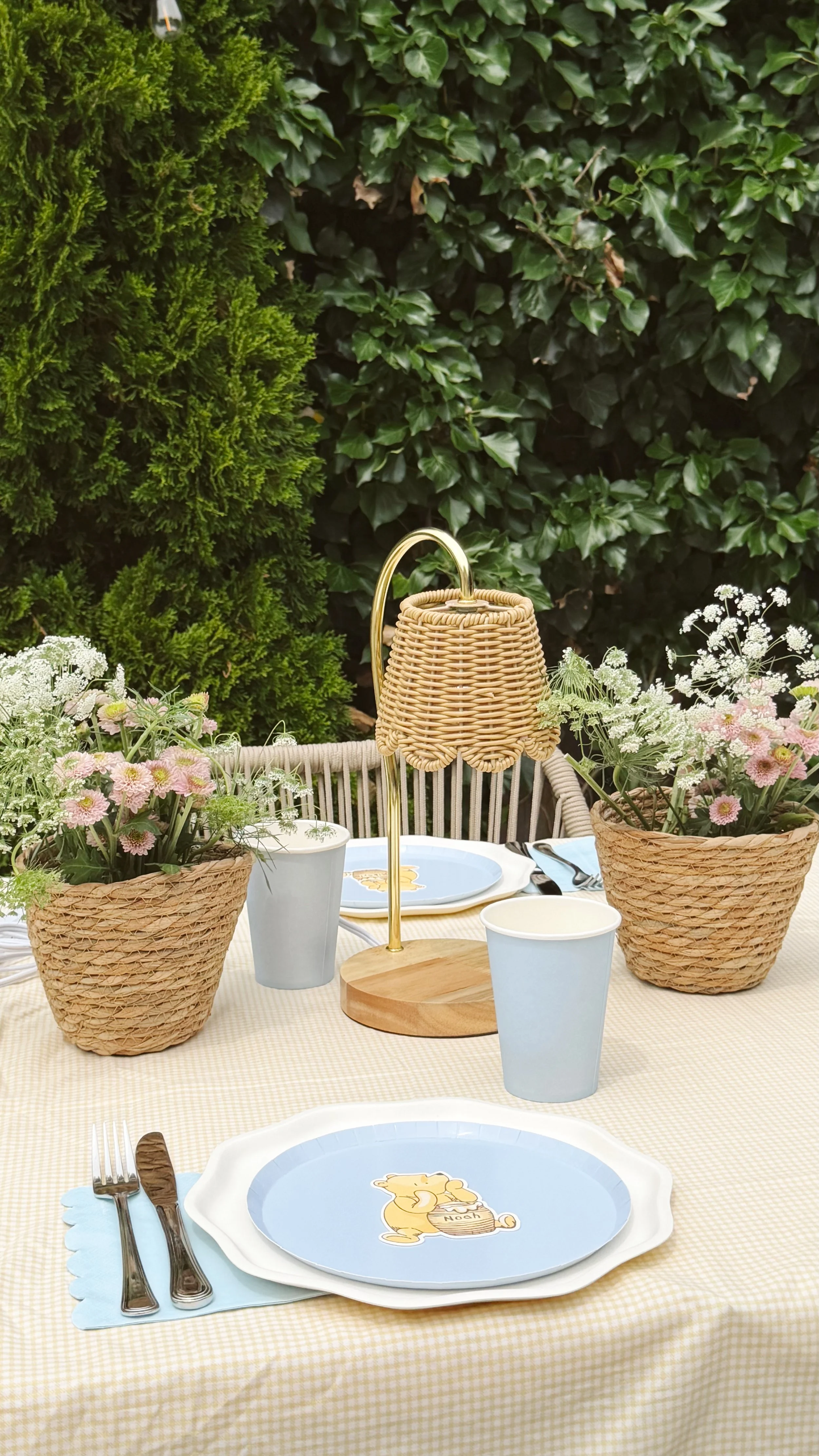 A table set outdoors with a yellow checkered tablecloth, containing a white plate with a cartoon image of a teddy bear, three light blue cups, silverware, and a napkin. There are two woven baskets with pink and white flowers, a small white cup with greenery, and a lamp with a wicker shade. The background features green foliage.