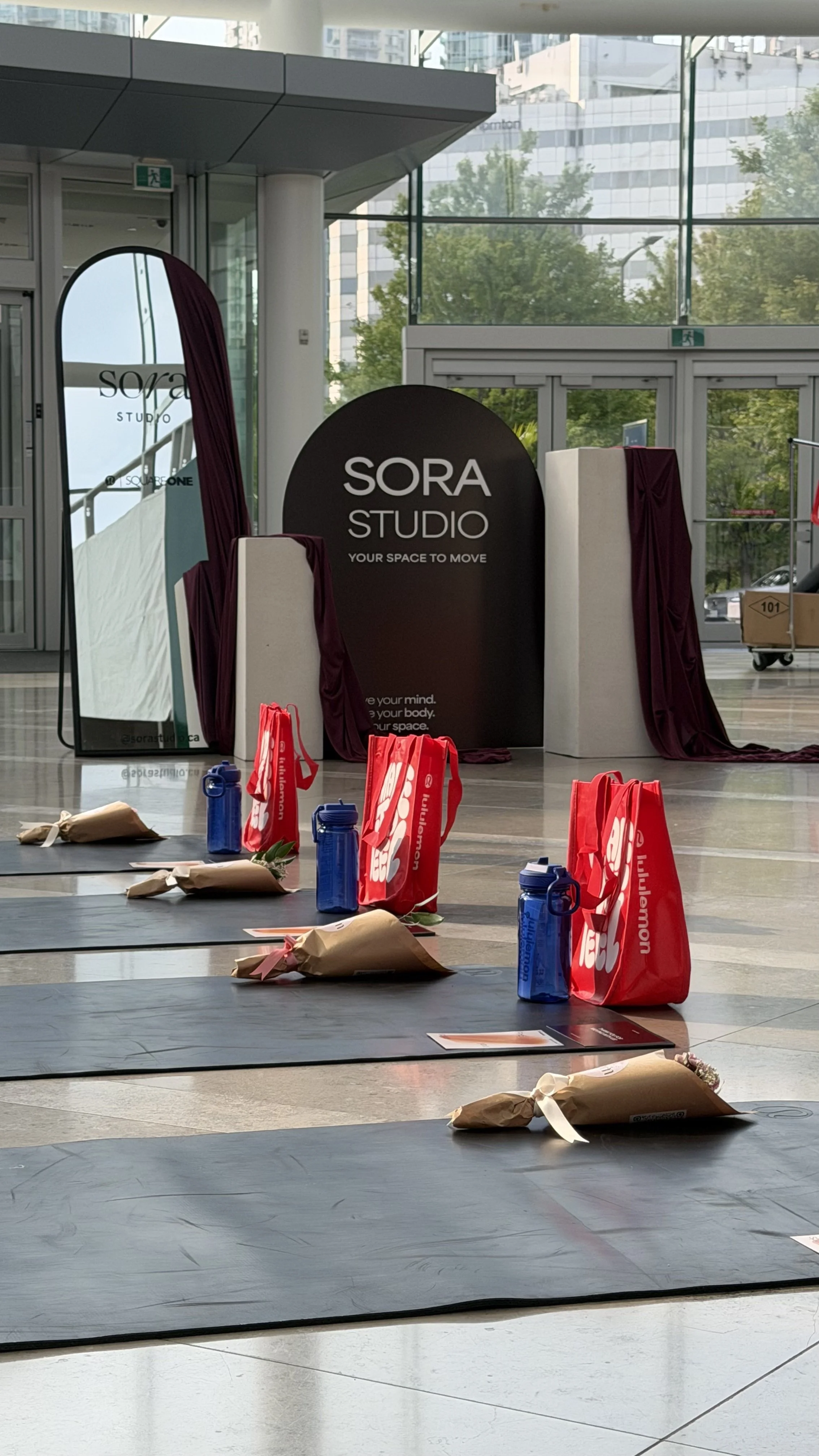 Inside a modern building, an event setup with mats and gifts arranged on the floor, a mirror, and a sign that reads 'SORA STUDIO' with the tagline 'Your space to move,' large windows showing trees and city buildings outside.