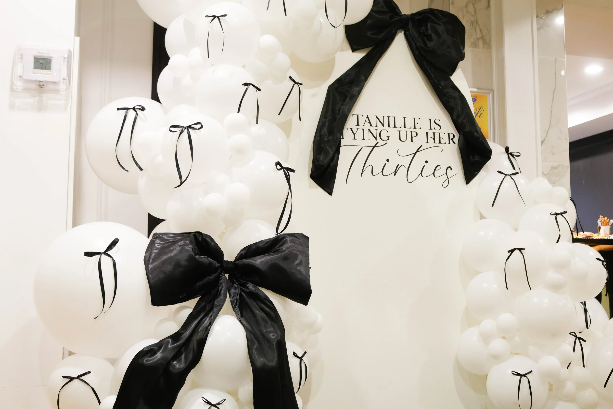 A decorative display with numerous white balloons, each adorned with a small black bow, surrounding a black and white sign that reads, 'TANILLE IS TYING UP HER THIRTIES.' The sign is decorated with a large black satin bow at the top. The background a
