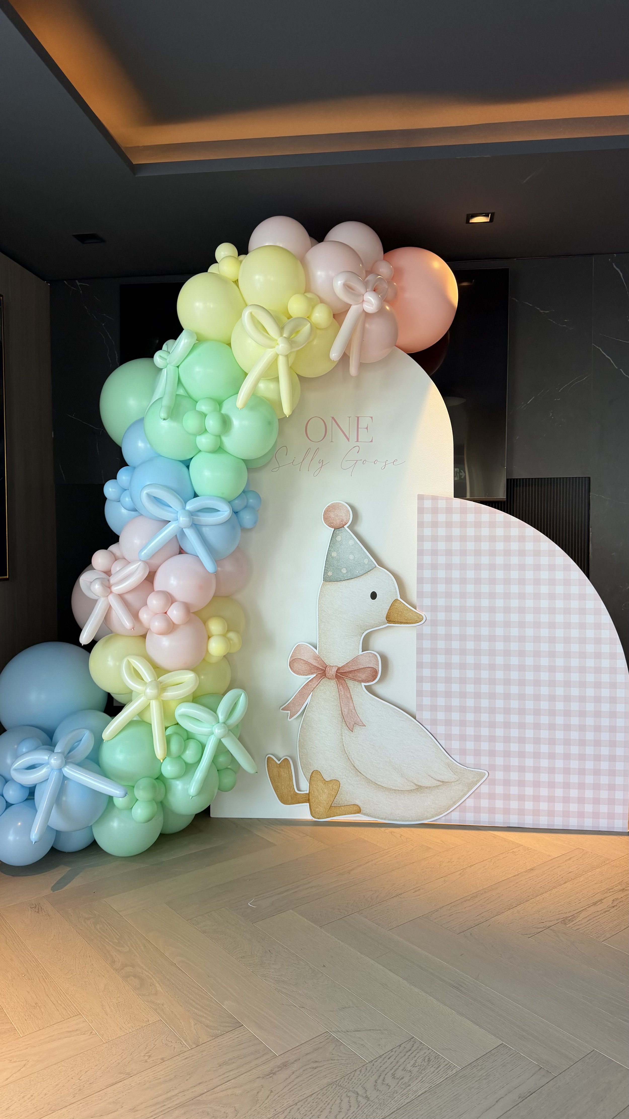 Decorative display with pastel-colored balloons arranged to resemble a bunch of grapes, with balloon bows. A sign with a cartoon goose wearing a party hat and bow, and text that reads 'One Silly Goose' is attached to a pink and white checkered backgr