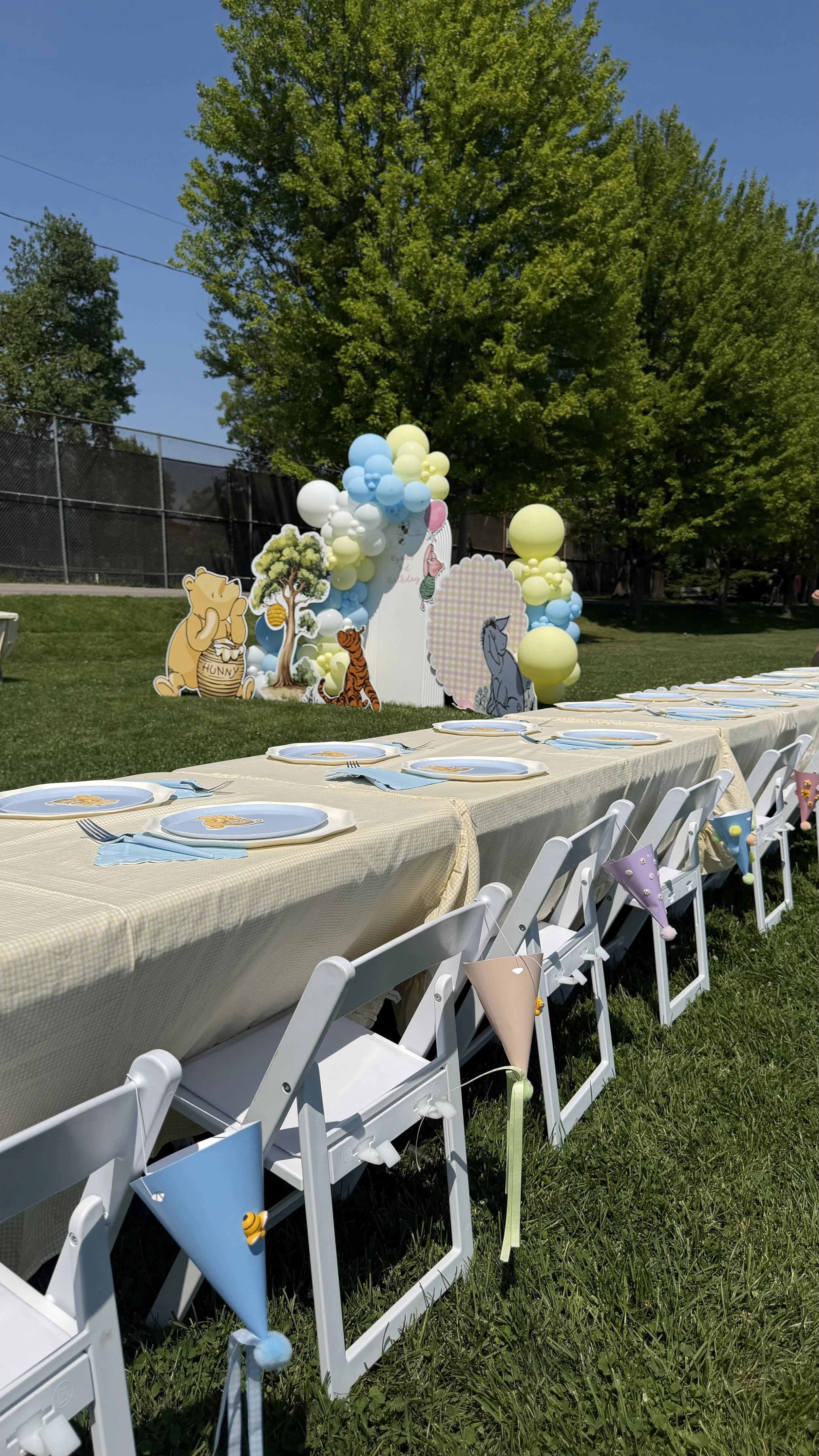 Outdoor birthday party setup with a long table covered with a yellow tablecloth, decorated with paper fans and bunting, and cartoon character cutouts from Winnie the Pooh, Tigger, and Eeyore, with a backdrop of balloons and trees under a clear blue s