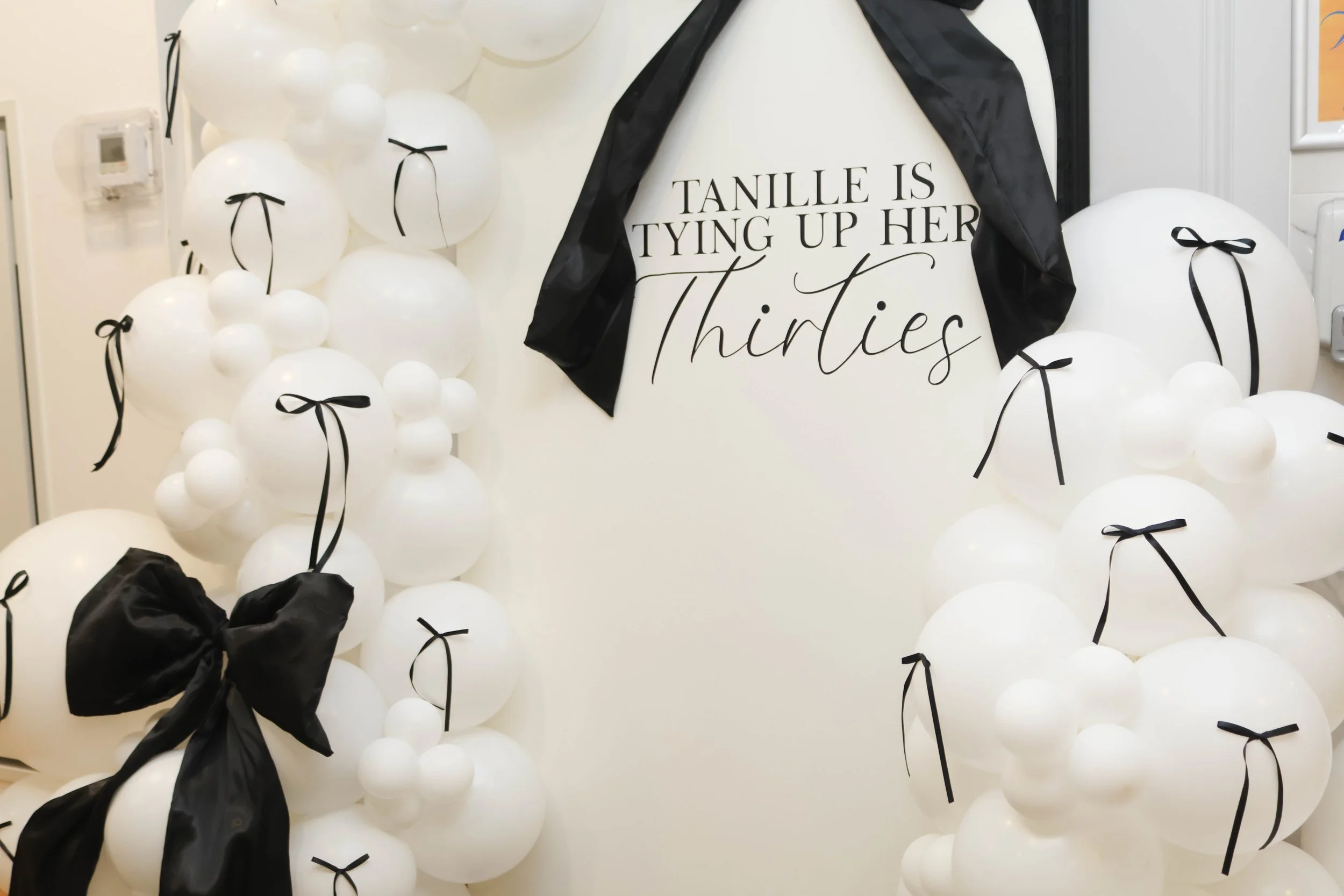 Wall decorated with white balloons tied with black ribbons and a sign reading 'TANILLE IS TYING UP HER Thirties' with black fabric draped around it.
