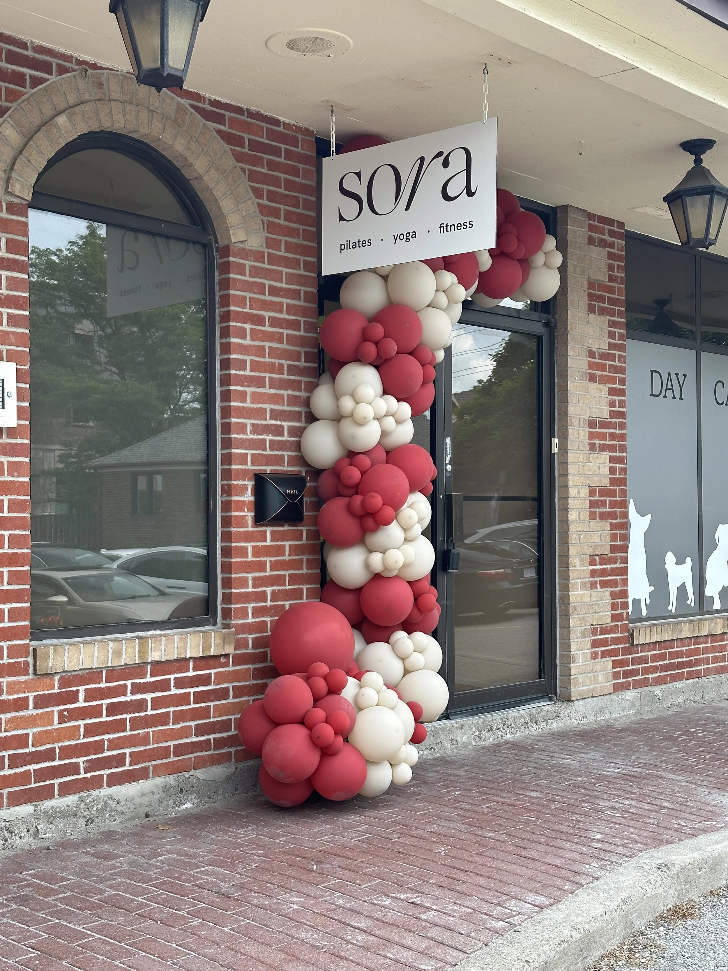 Storefront with a white sign reading 'sora' and smaller text for pilates, yoga, and fitness. Decorated with red and white balloons cascading beside the entrance, which is framed by a brick wall and large windows. There are lamps hanging from the ceil