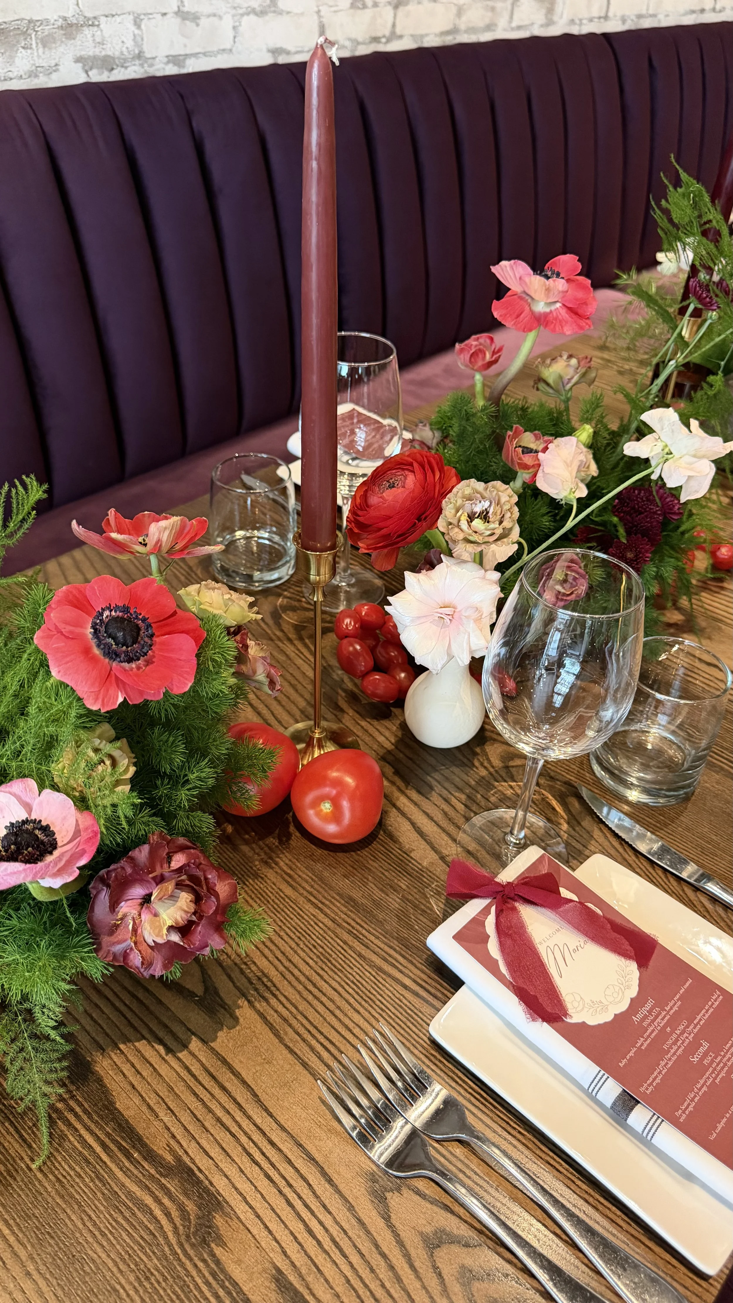 A decorated dining table with pink and red flowers, a pink candle, empty wine glasses, a menu with a ribbon, and fresh tomatoes on a wooden table against a brick wall.