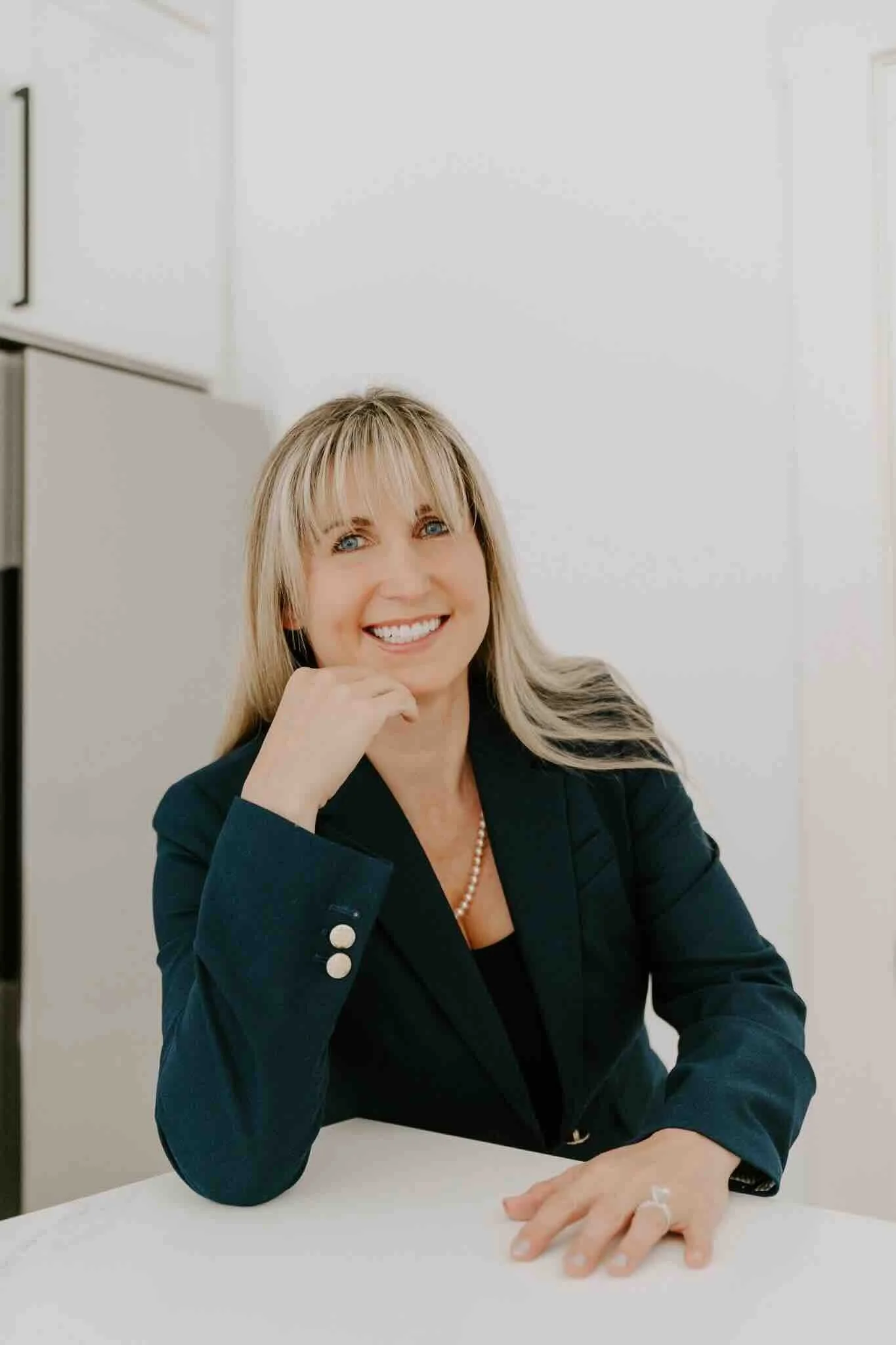 A woman with blonde hair and blue eyes smiling, resting her chin on her hand, seated at a white table in an office with white walls and cabinets.