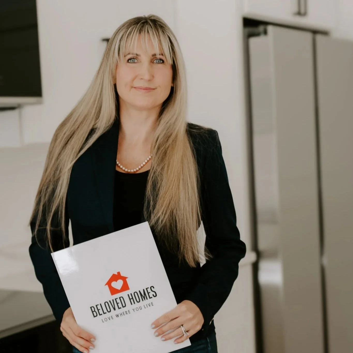 A woman with long blonde hair and blue eyes holding a booklet with a logo of a house with a heart inside it. She is wearing a black blazer, a pearl necklace, and a ring on her left hand. She is standing in front of a white wall with a cabinet and a television in the background.