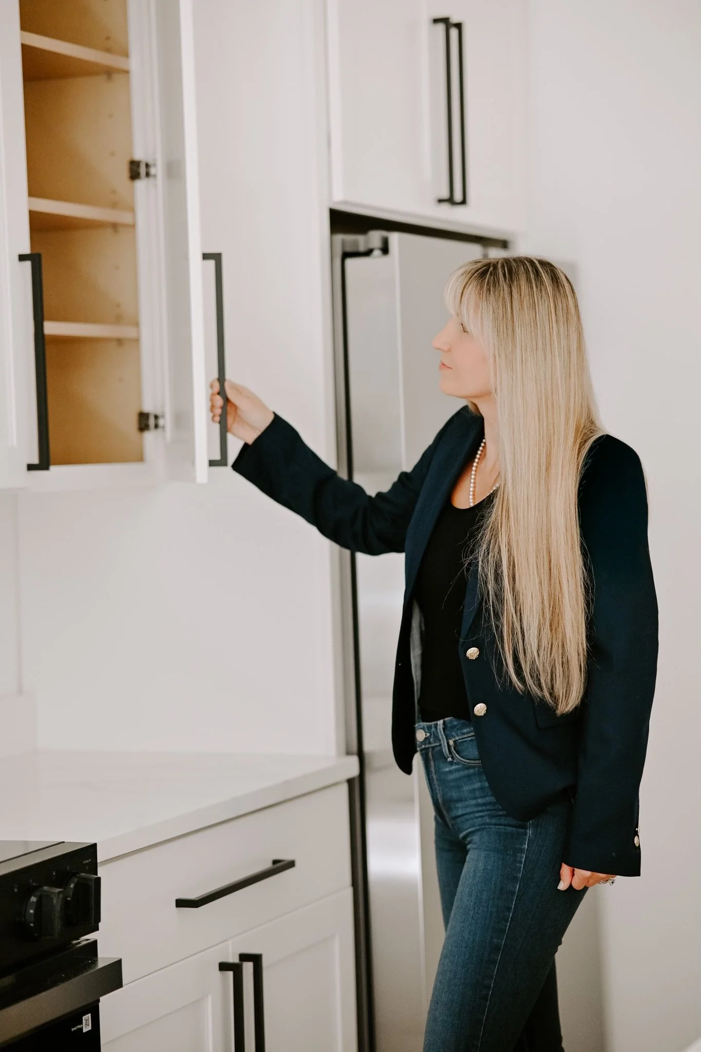 A woman with long blonde hair wearing a black blazer, black top, and jeans, looking at a white cabinet door in a modern kitchen.