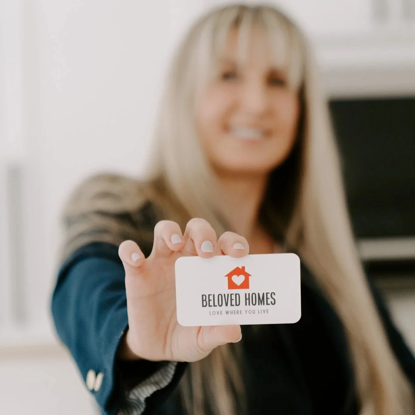 A woman with blonde hair holding a business card that reads 'Beloved Homes' with a house and heart logo. The background is blurred.
