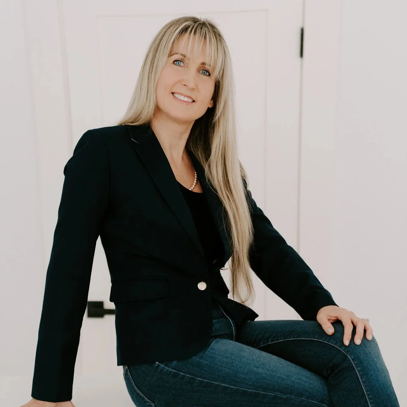 A woman with long blond hair, wearing a black blazer, black top, and blue jeans, sitting on a table in a white room, smiling at the camera.
