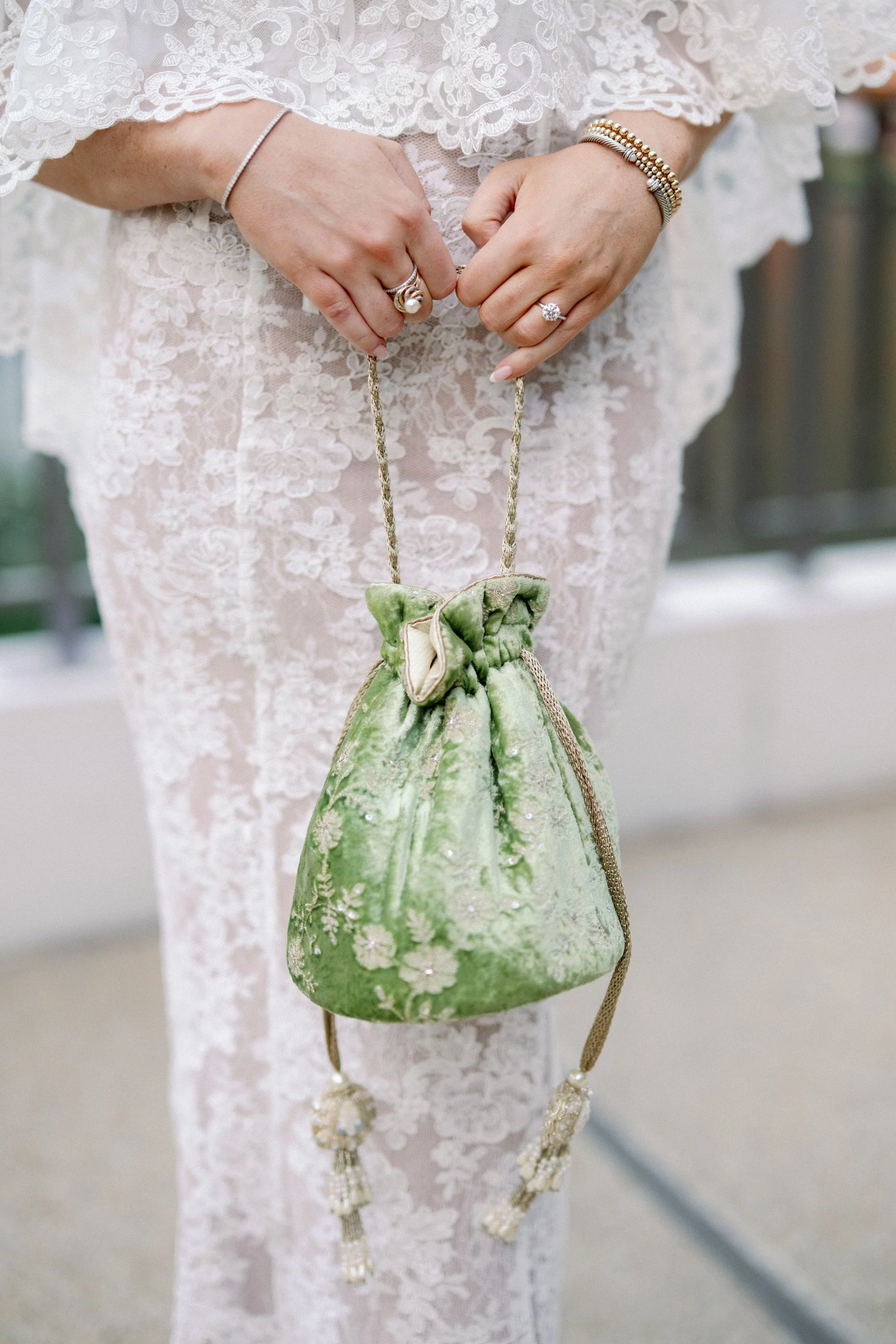 Close-up of a person holding a small, green velvet drawstring purse with a bow, standing in front of white lace fabric, wearing jewelry including rings, bracelets, and a thin bracelet, with additional blurred elements in the background.