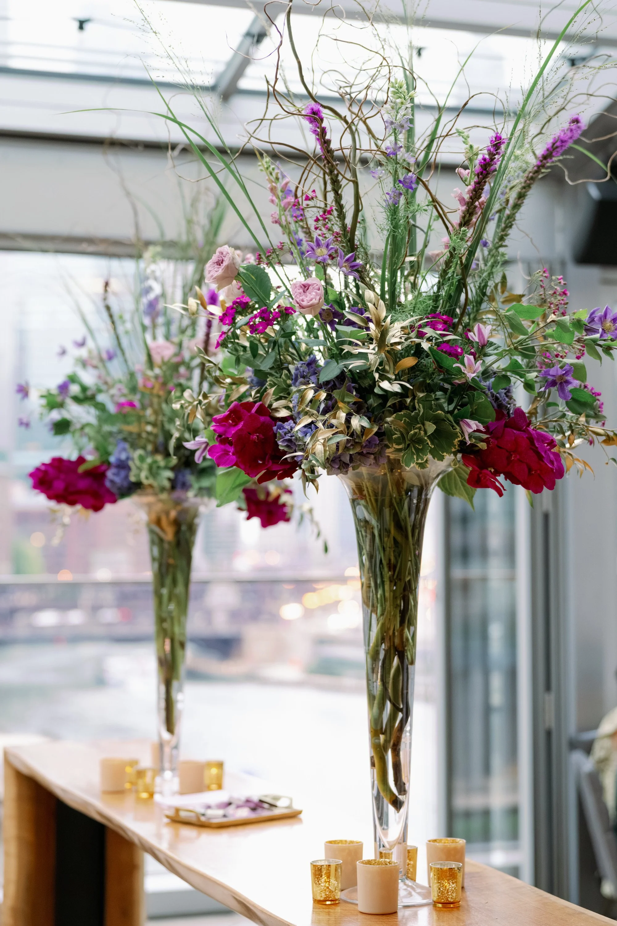 Elegant floral arrangement in tall vases with pink, purple, and red flowers, set on a wooden table with small gold and beige candles around the base, inside a bright room with large windows.