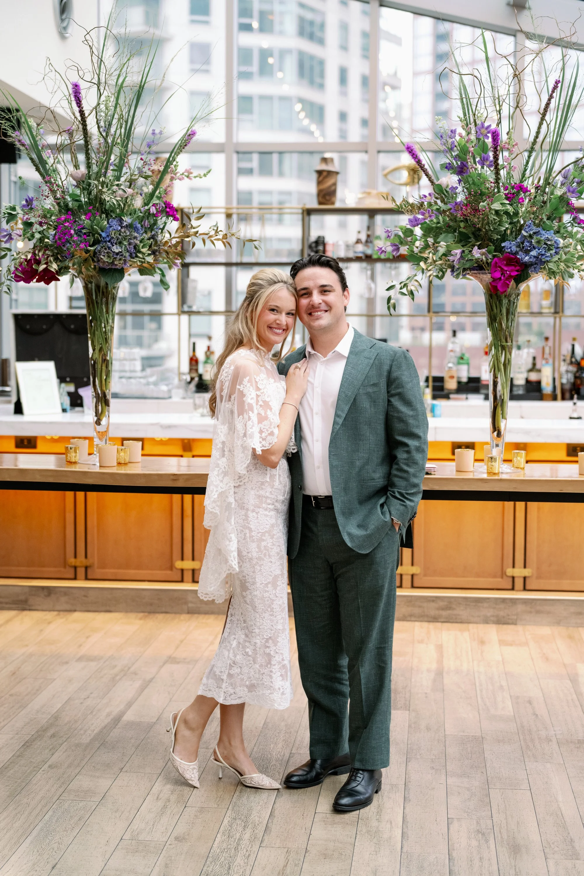 A smiling man and woman dressed in formal attire at an indoor event, standing in front of large floral arrangements on a bar counter. The woman is wearing a white lace dress and high heels, while the man is in a gray suit with a white shirt and black