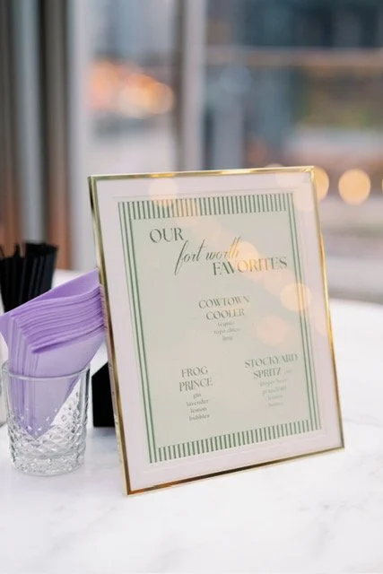 Framed sign listing favorite cocktail options, placed on a table next to a glass filled with purple napkins, with a window in the background.