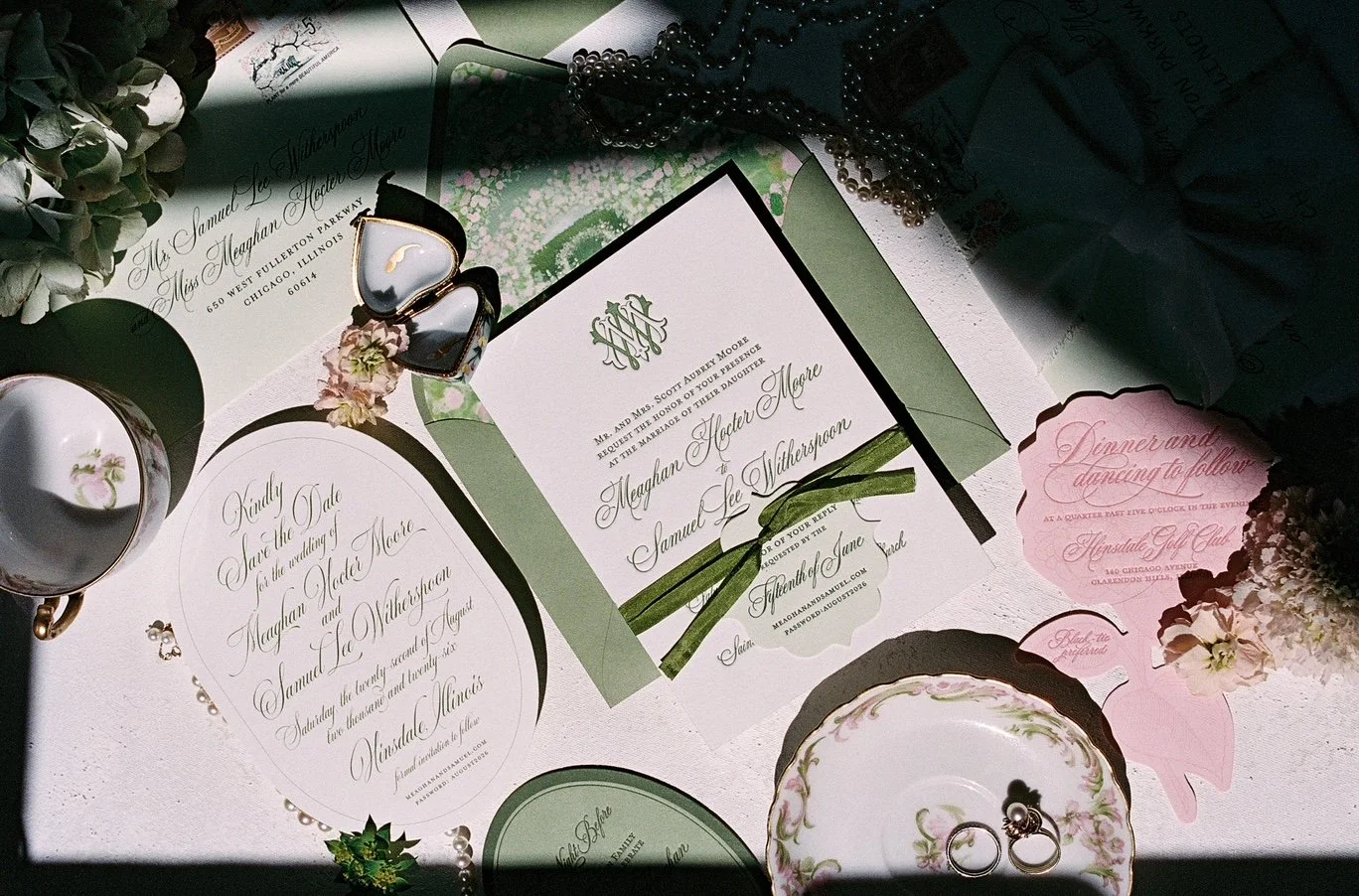 Wedding invitation suite with pink and green accents, including invitations, RSVP card, and envelopes, surrounded by jewelry, glasses, pearls, and flowers on a white surface.