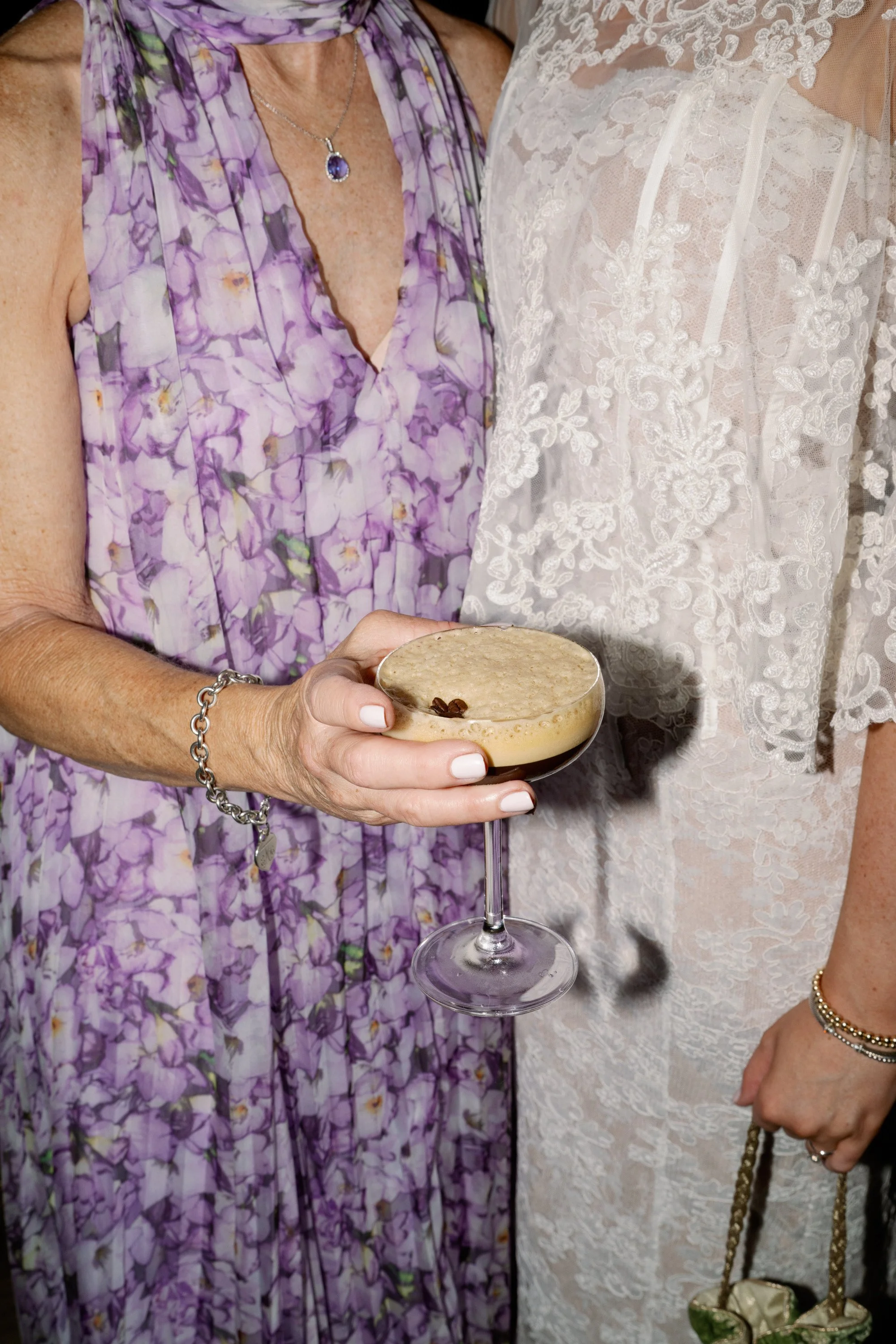 Close-up of two women dressed in elegant attire, one in a purple floral dress holding a cocktail glass with a creamy beverage garnished with a coffee bean, and the other in a white lace dress holding a handbag with a chain strap.