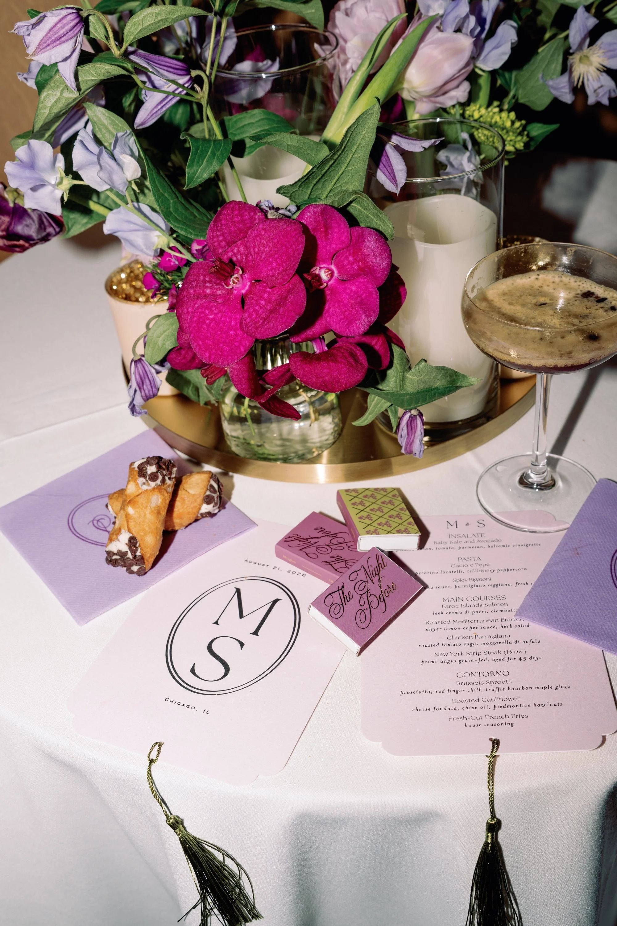 A floral centerpiece with pink orchids, purple sweet peas, and other flowers, accompanied by candles in glass holders, on a table with menus and a dessert plate.