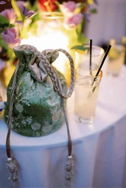 A decorative drawstring bag with a floral pattern and two glasses of a light-colored beverage with black straws on a table, with flowers in the background.