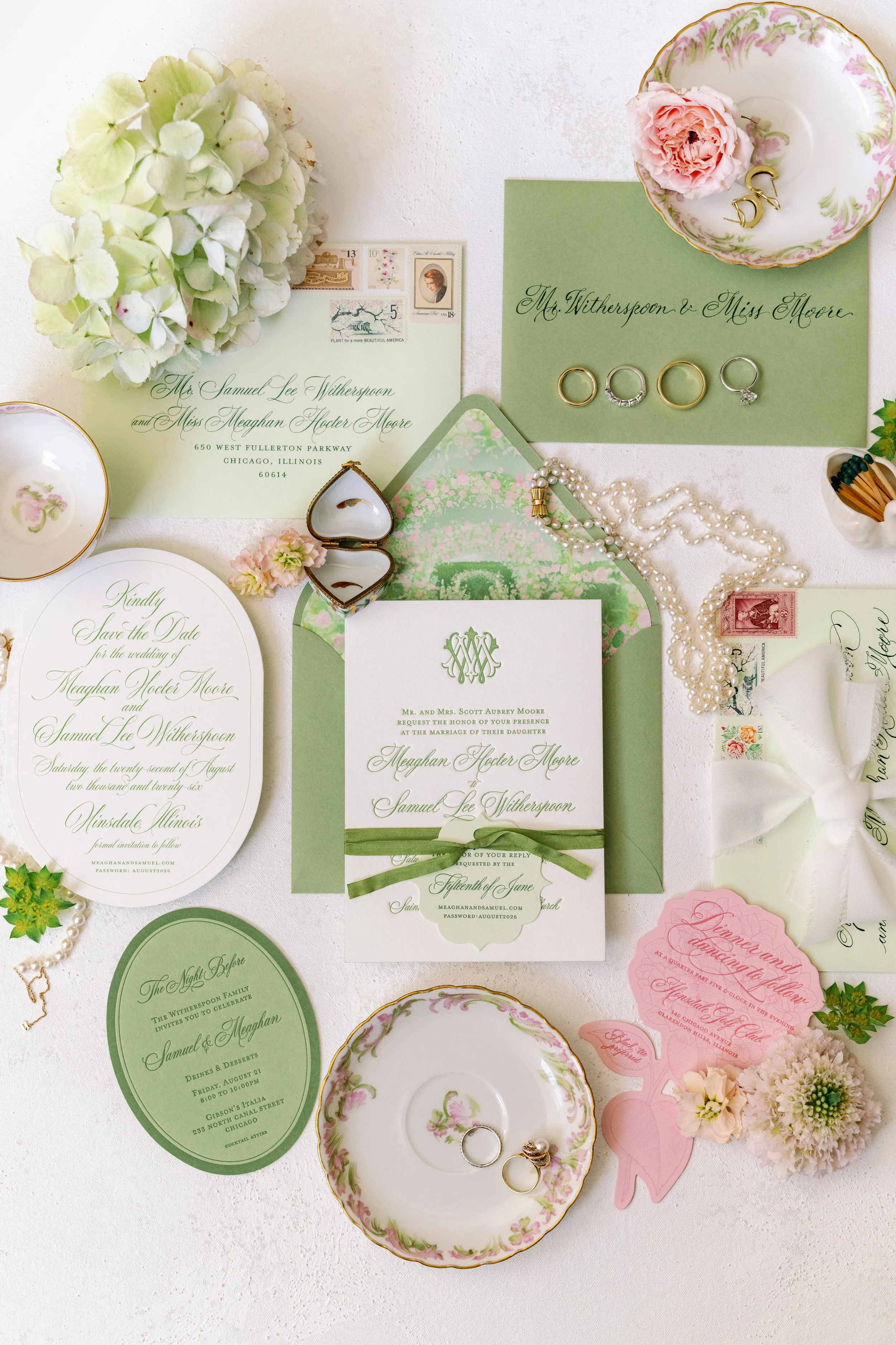 An assortment of wedding invitations, rings, peonies, and decorations arranged on a white surface, featuring vintage floral china and accented with pearls and pink hair accessories.