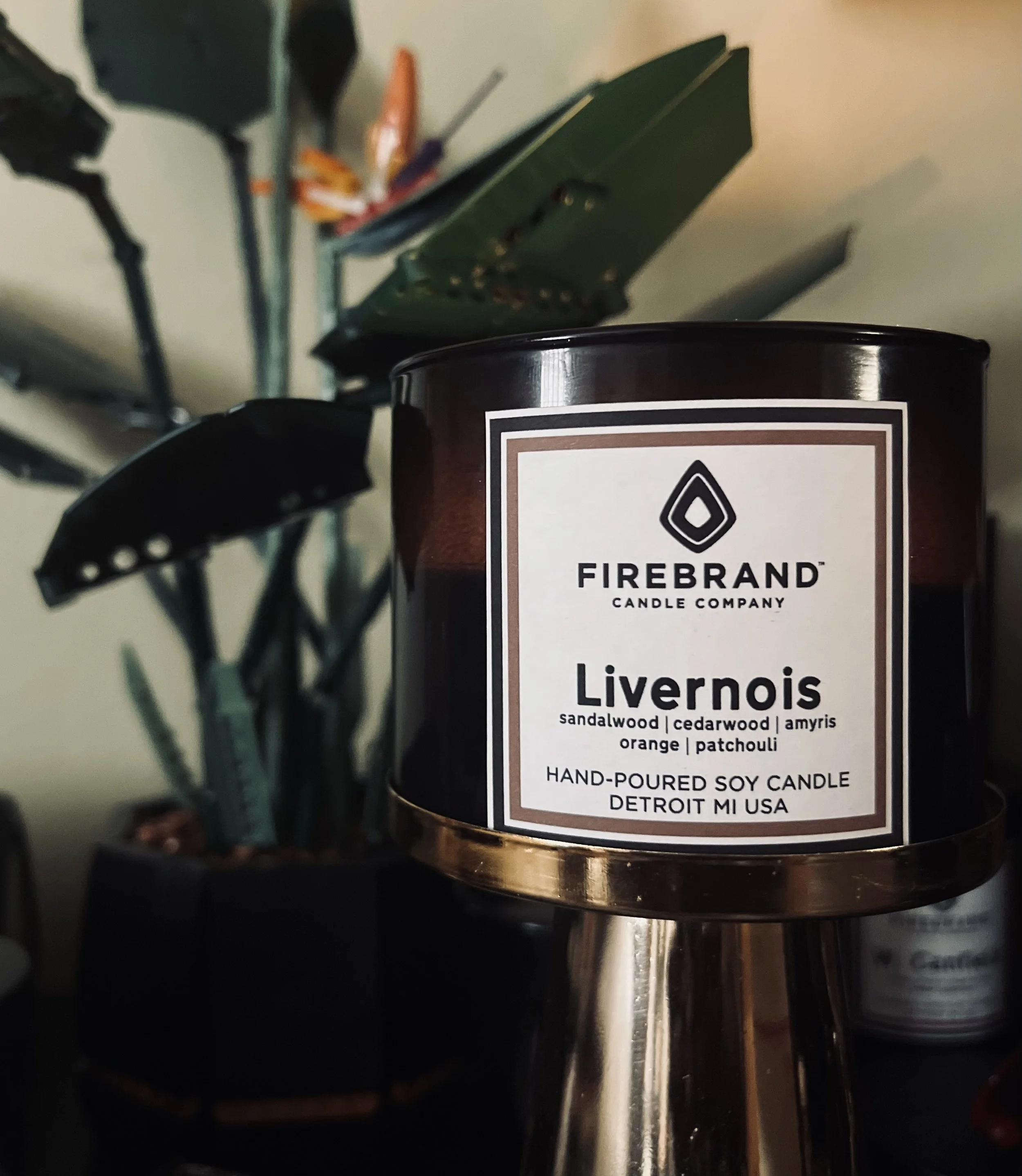 Close-up of a candle with a white and black label from Firebrand Candle Company, highlighting the scent 'Livernoise' which includes sandalwood, cedarwood, amyris, orange, and patchouli. The background shows gardening tools hanging on a wall, slightly out of focus.