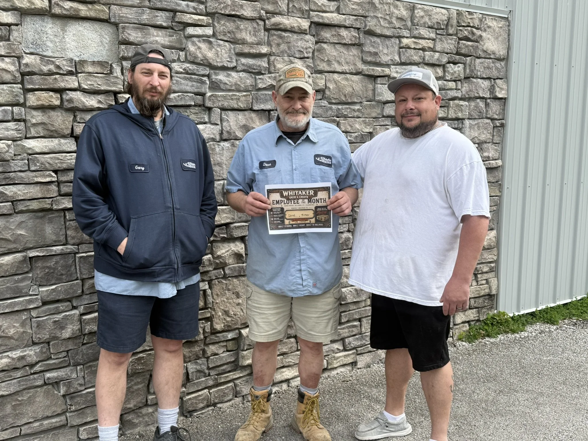 Three men standing outdoors in front of a stone wall. The man in the middle is holding a framed award for Employee of the Month. The man on the left is wearing a blue hoodie and cap, the man in the middle is wearing a light blue shirt and a cap, and the man on the right is wearing a white t-shirt and a cap. They are all smiling and looking at the camera.