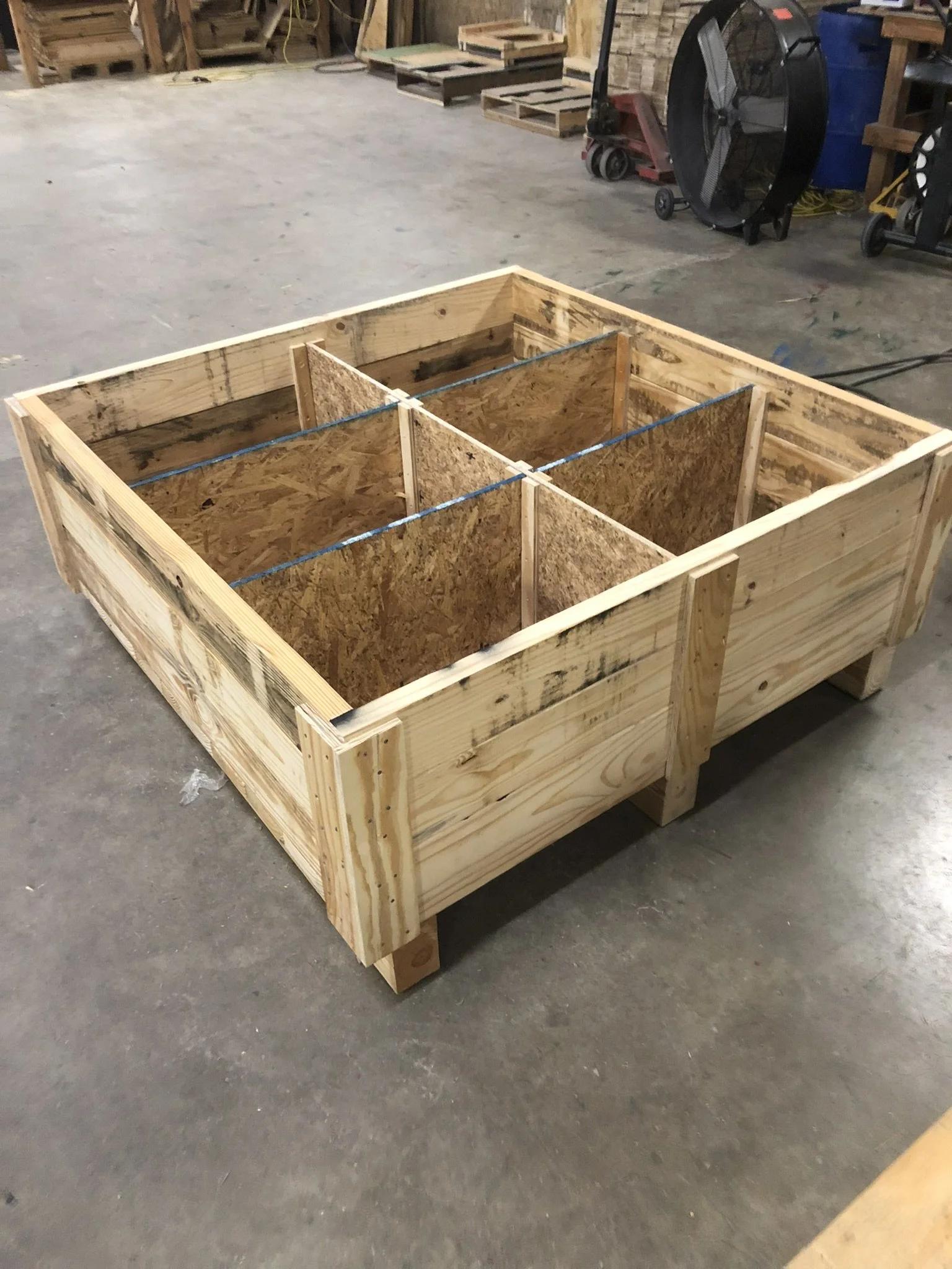 A wooden construction frame in a workshop, with plywood sides and a divider, supported by wooden blocks on a concrete floor.
