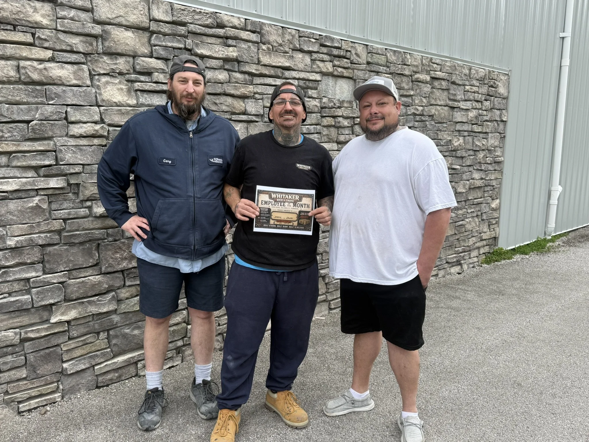 Three men standing outdoors in front of a stone wall and a metal building, one holding a framed certificate that reads 'Employee of the Month.' They are wearing casual clothing and hats, smiling at the camera.