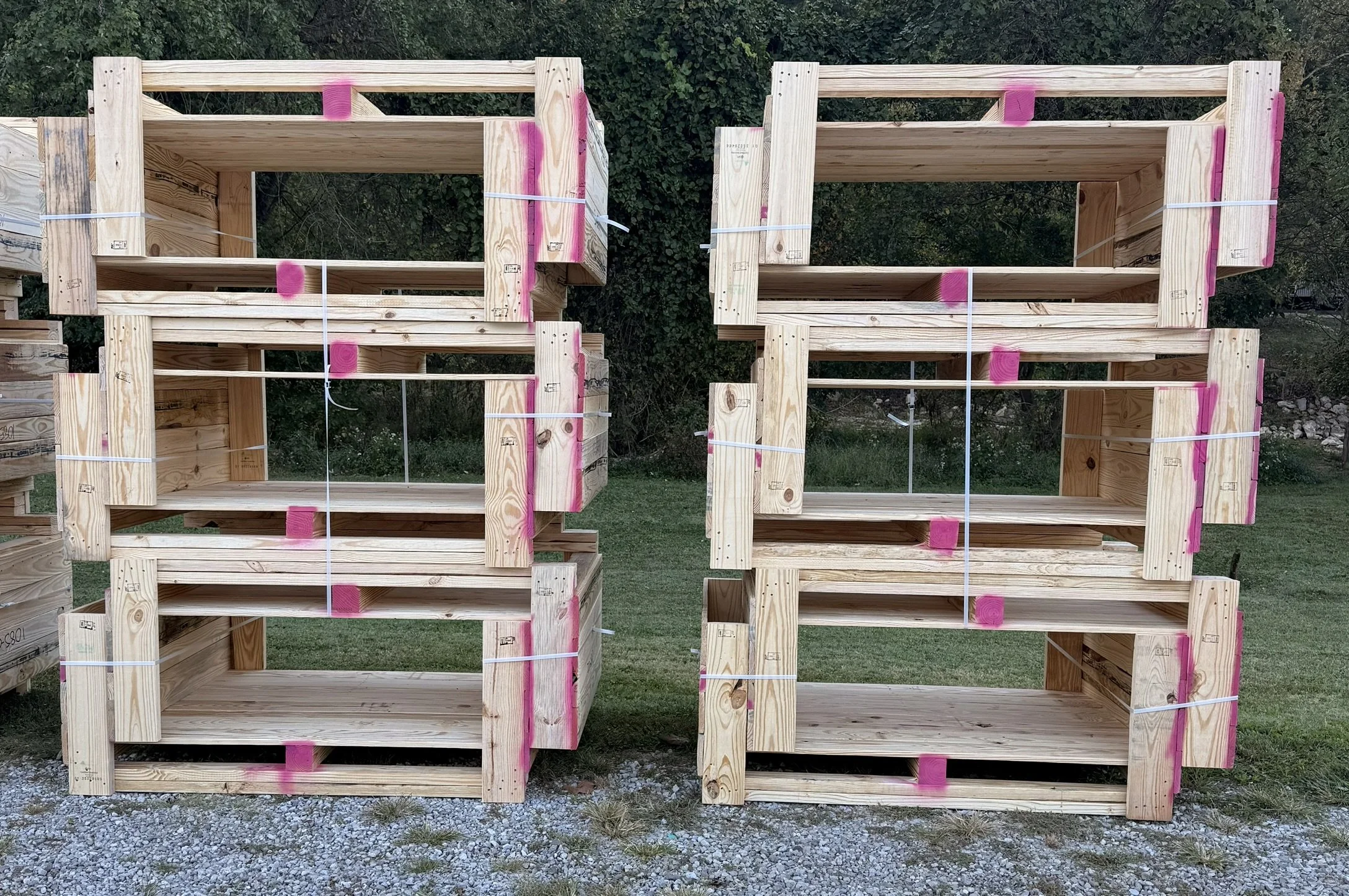 Construction site with stacked wooden pallets marked with pink spray paint, placed outdoors on gravel with trees in the background.