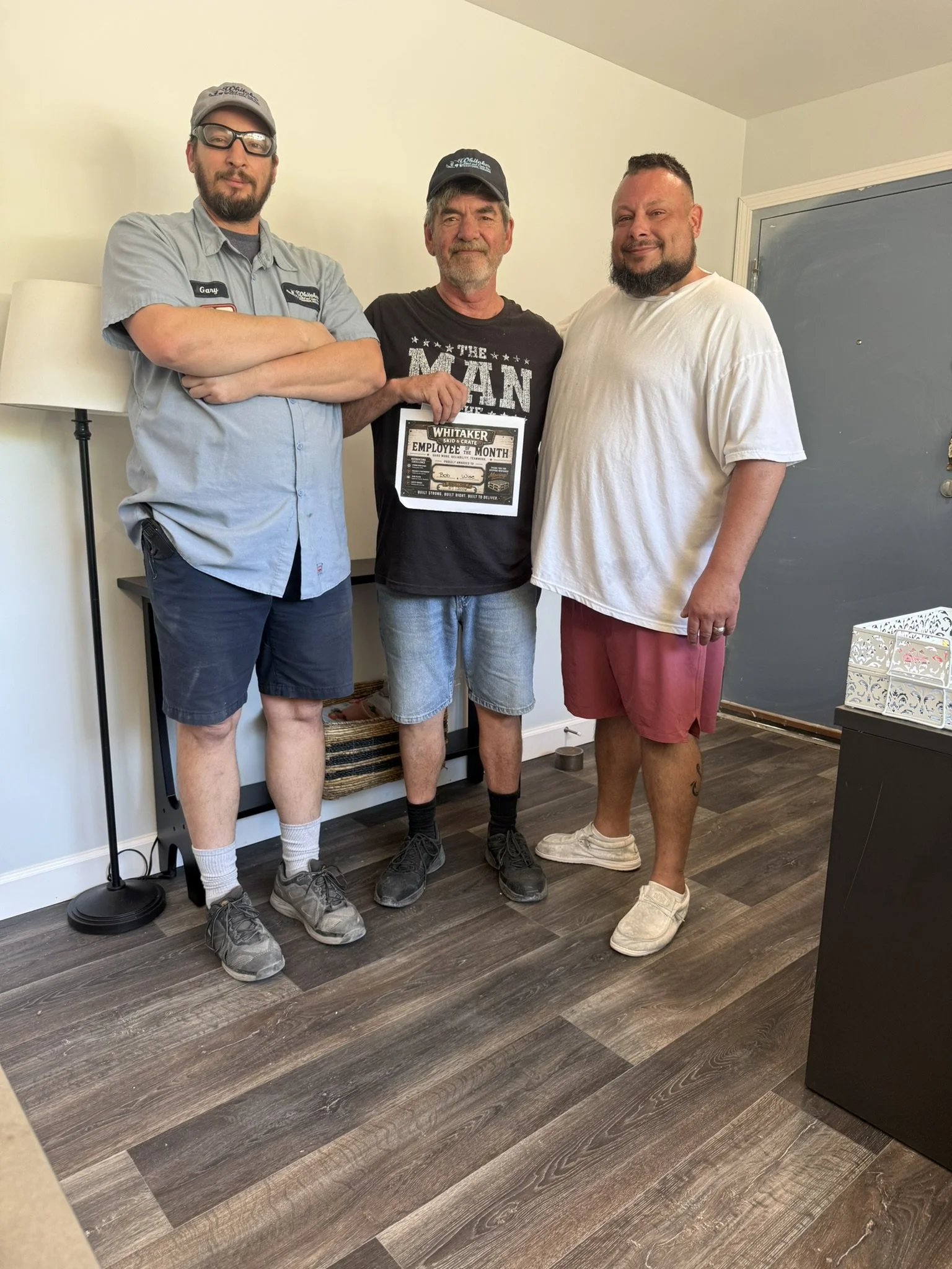 Three men standing together indoors, one holding an employee of the month certificate, all smiling or with neutral expressions, near a door and a lamp.